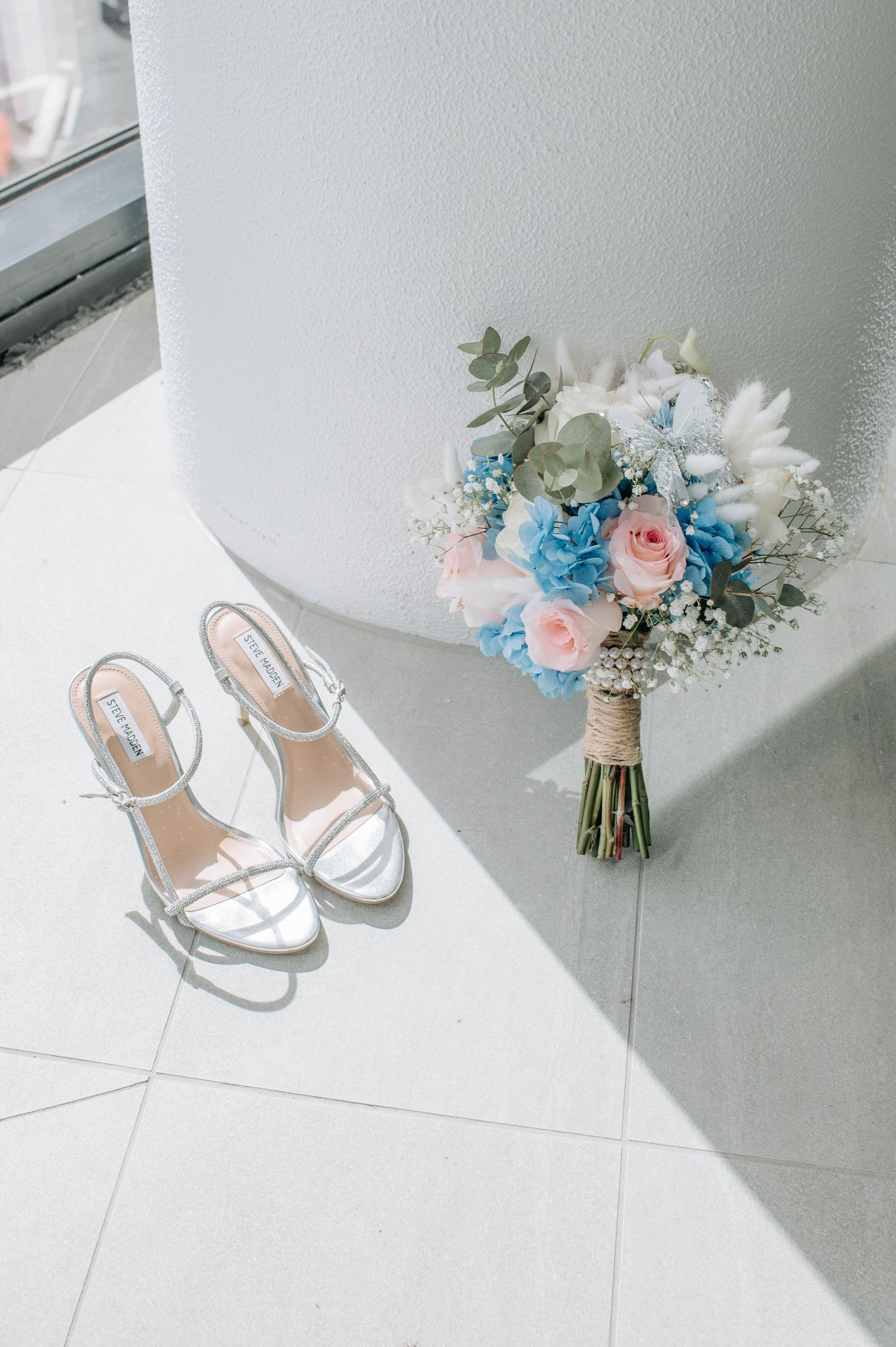 Wedding accessories including silver shoes and a blue and pink floral bouquet on white tile floor.