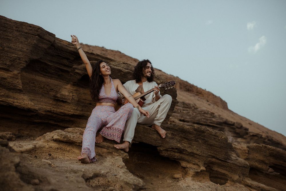 A couple embraces while sitting on rocky coastal cliffs overlooking the ocean at sunset.