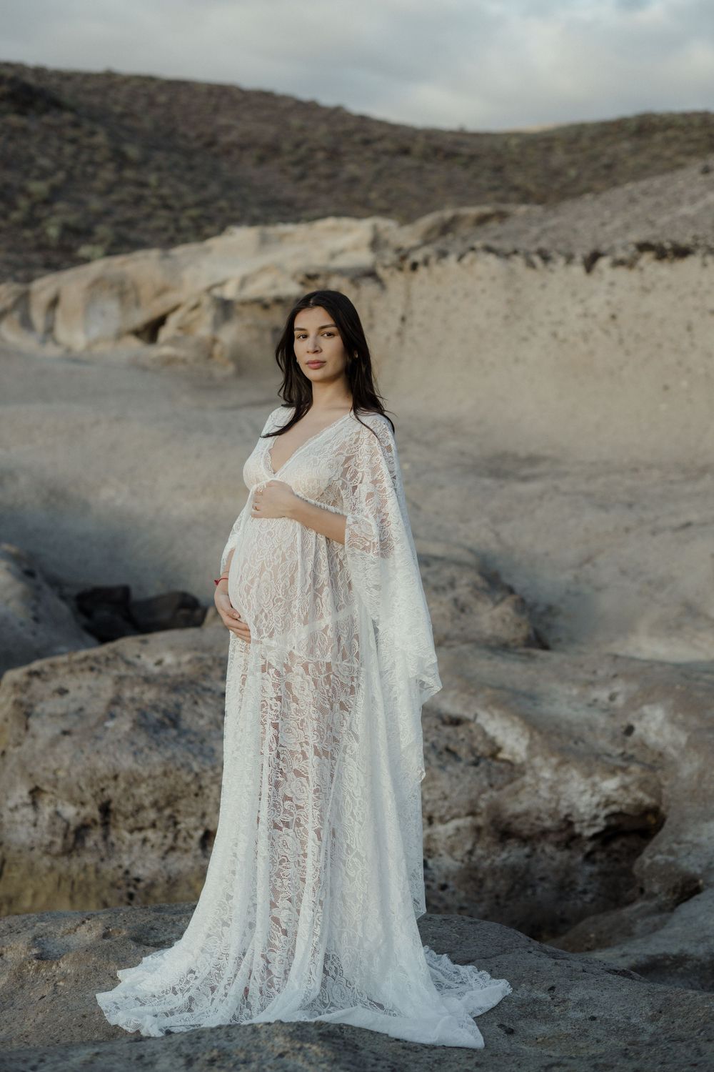A flowing white lace maternity gown with cape sleeves photographed against rocky desert terrain at sunset.