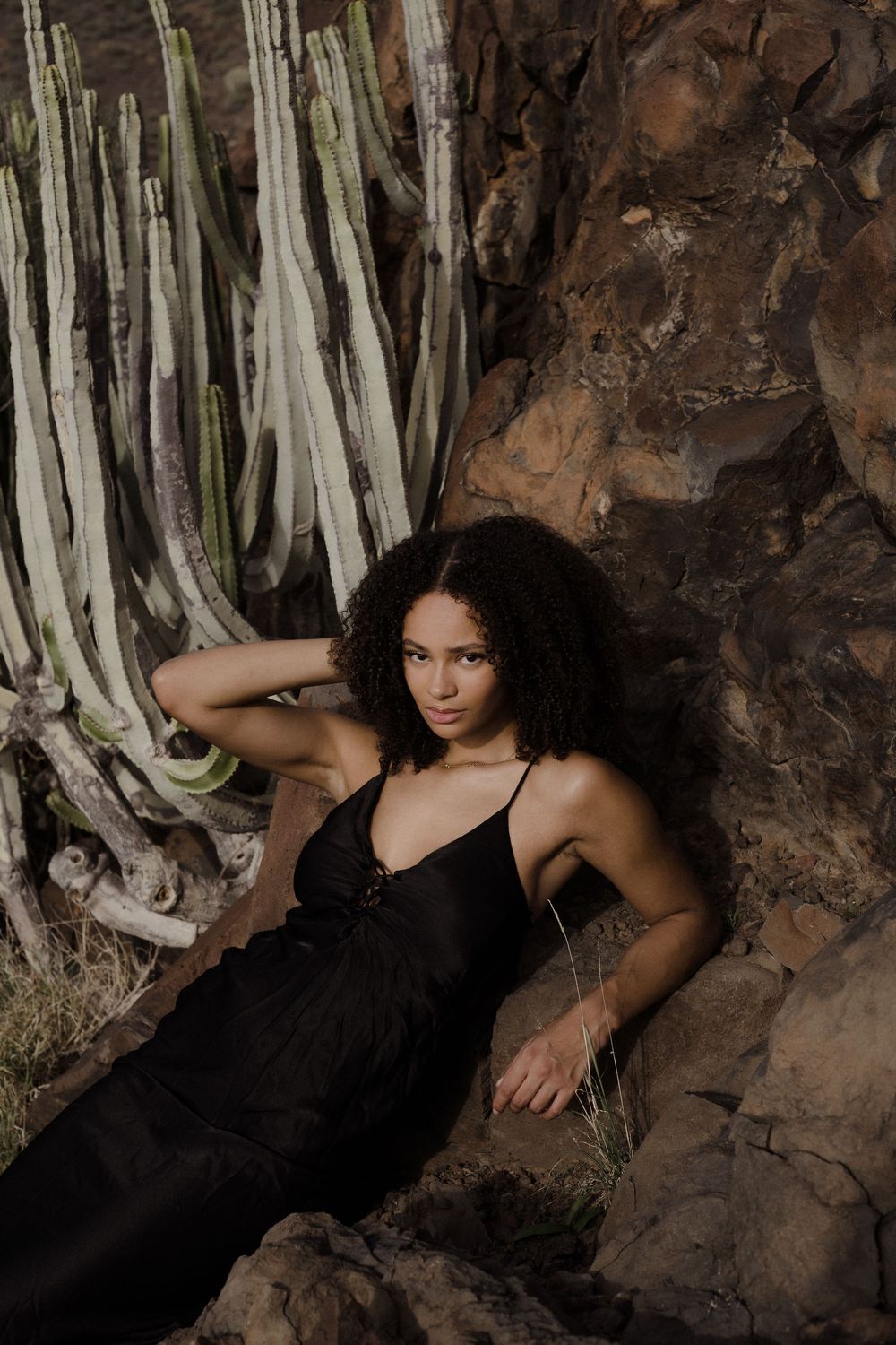 A person in a black dress poses dramatically against a rocky cave wall with hanging white plants in the background.