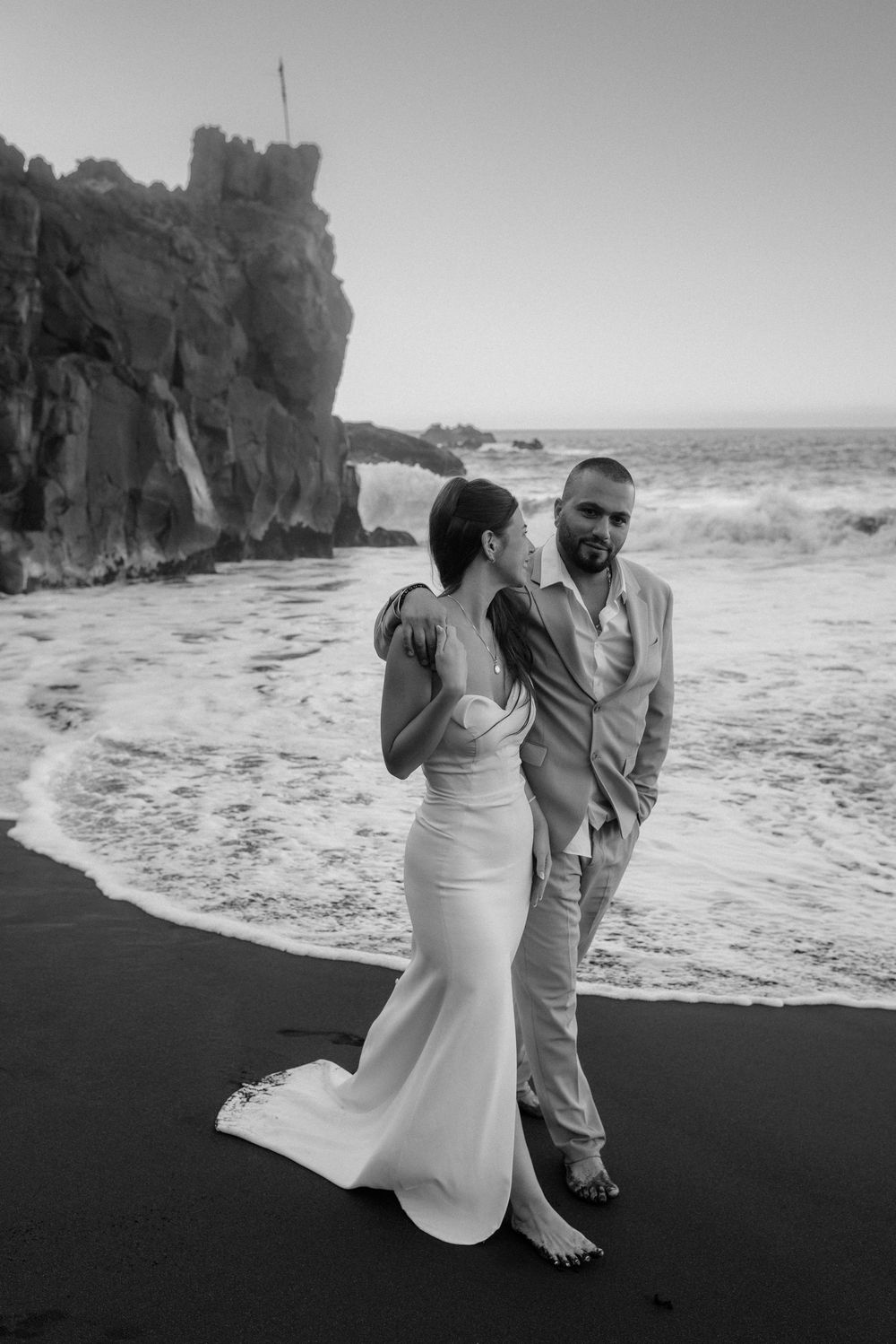 A couple shares an intimate moment on a black sand beach with rocky cliffs and ocean waves in the background.