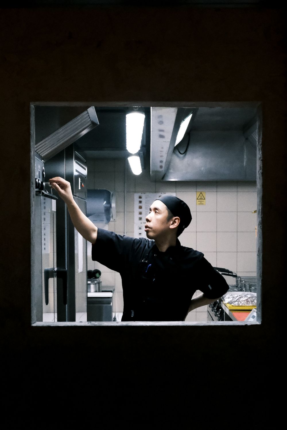 Illuminated kitchen service window showing worker in dark uniform reaching up.