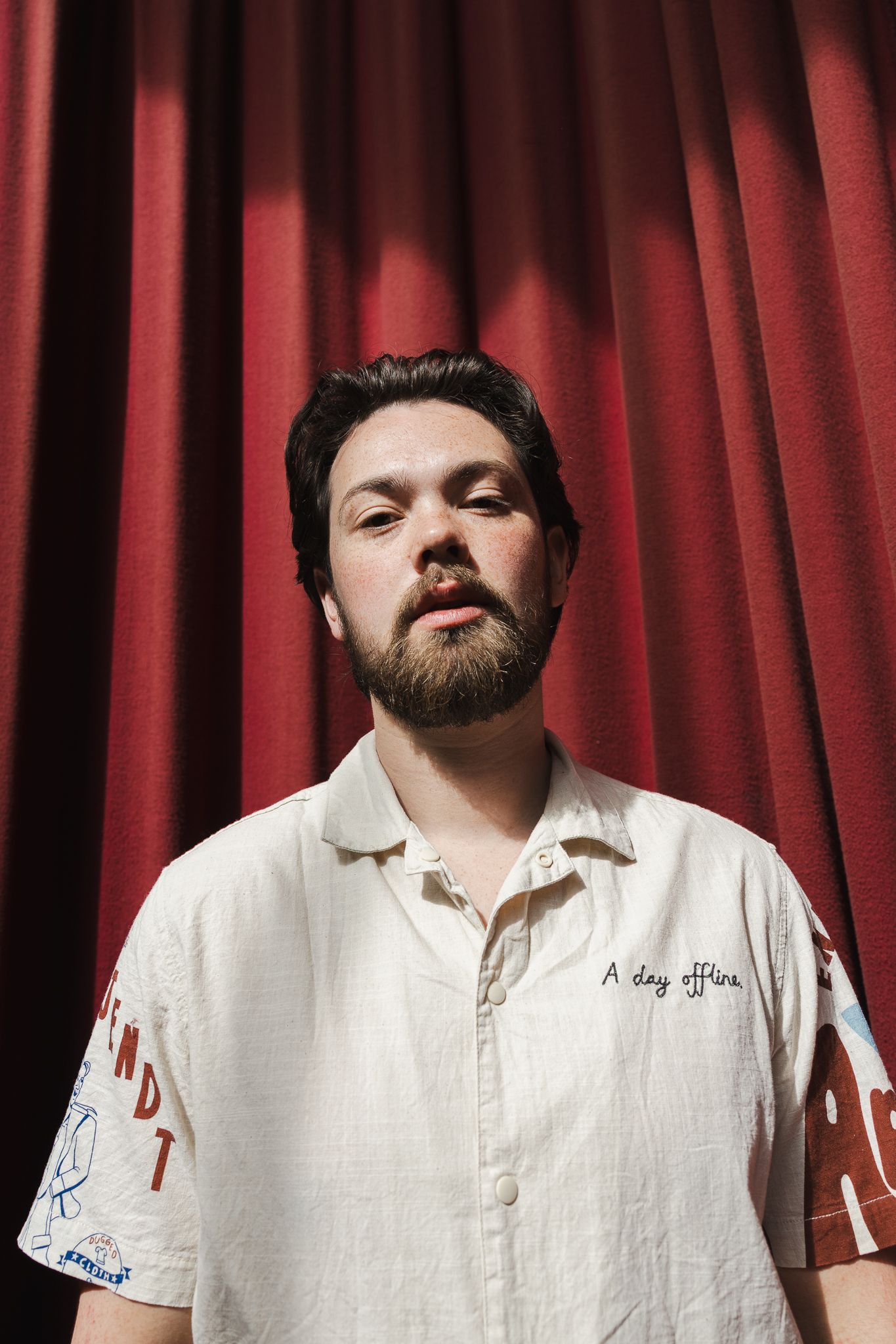 A bearded man in a white shirt stands before a red curtain backdrop in a portrait photo.
