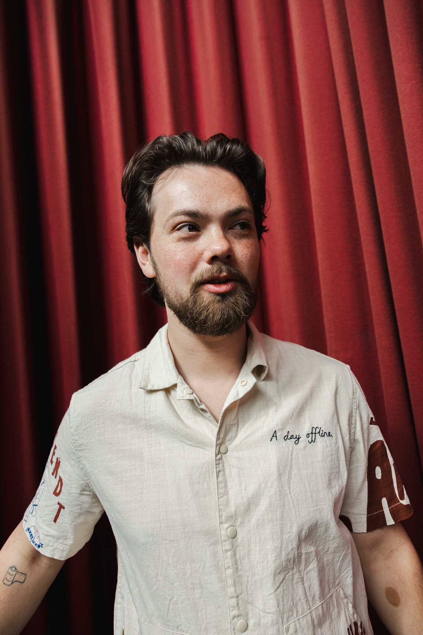 Bearded man in cream button-up shirt stands before red curtain, looking sideways with tattoos visible on arm.