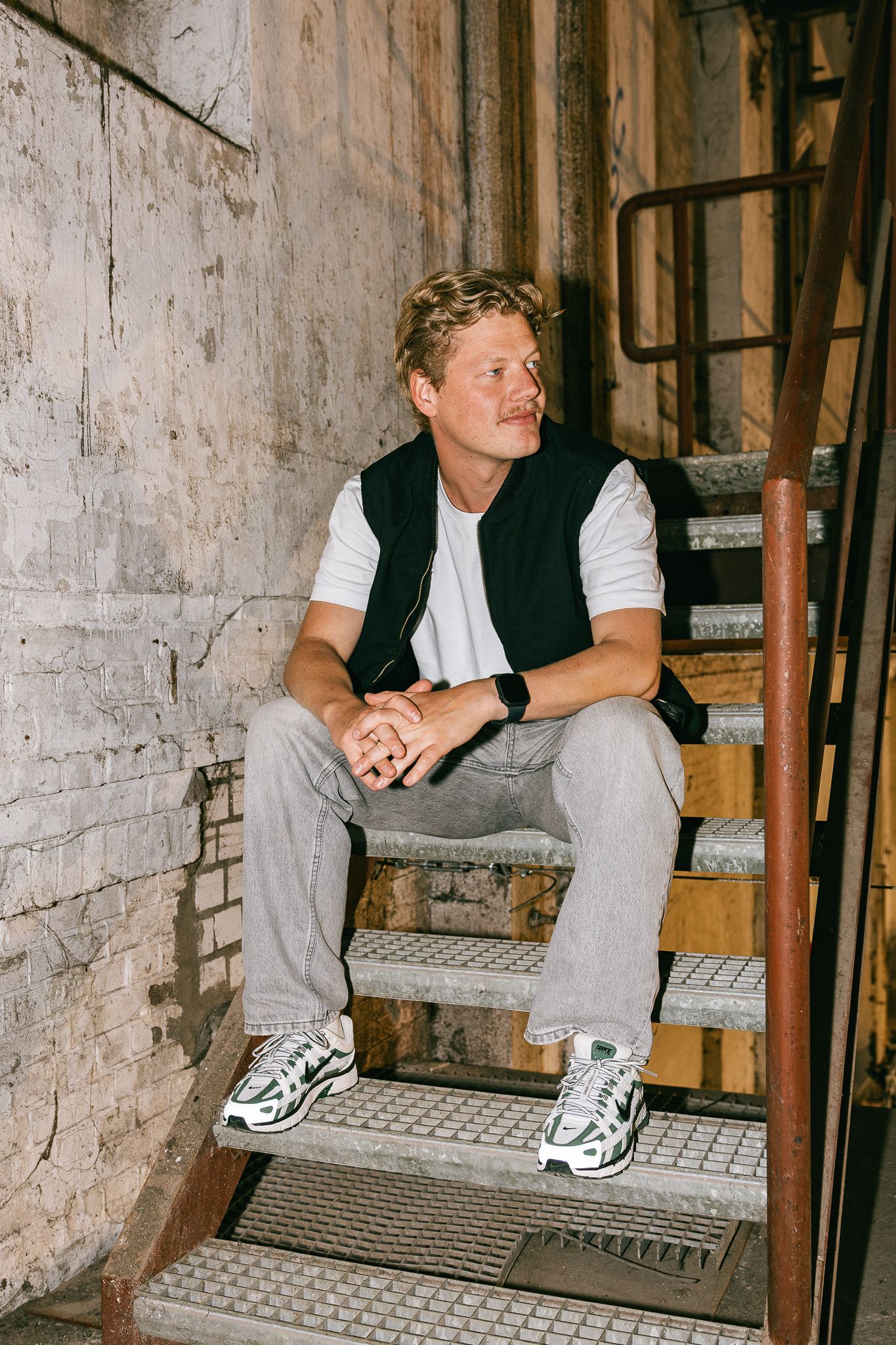 Person wearing a black vest and white t-shirt sits casually on industrial metal stairs with brick wall backdrop.