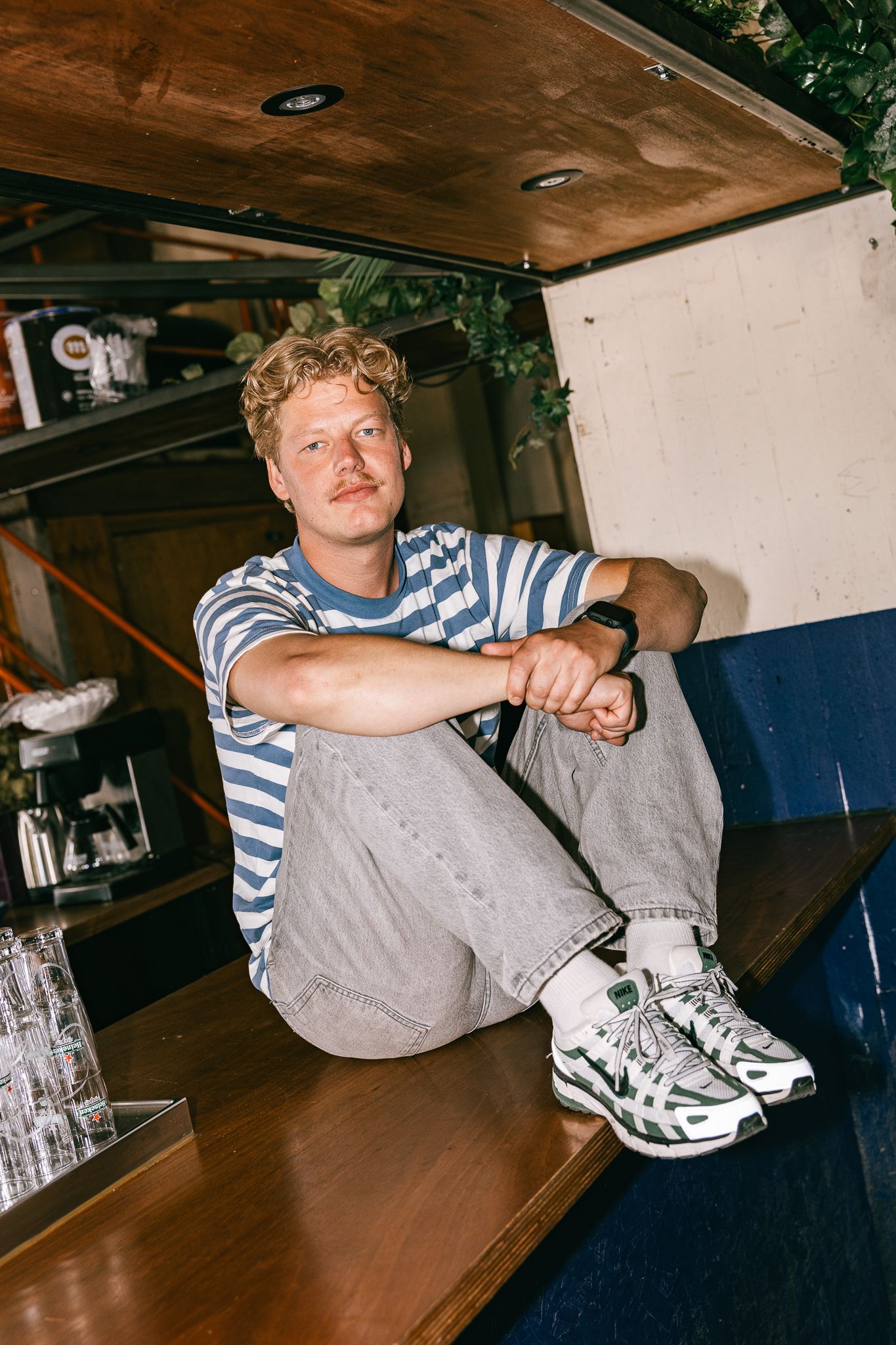 Person in striped shirt relaxes on wooden bench with blue cushions wearing chunky white sneakers in casual indoor setting.