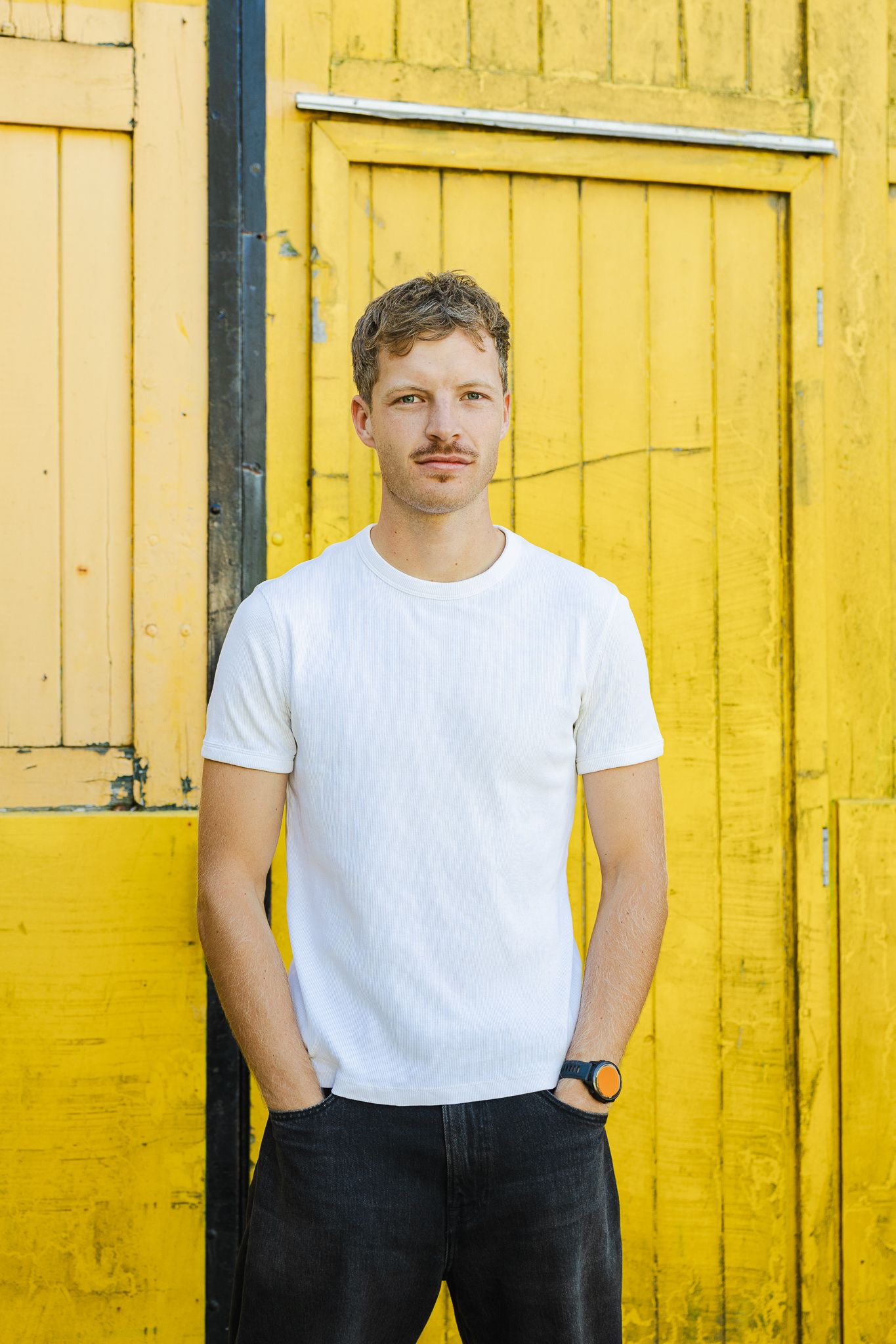 Young man with mustache in white t-shirt and black jeans standing against a vibrant yellow wooden door.