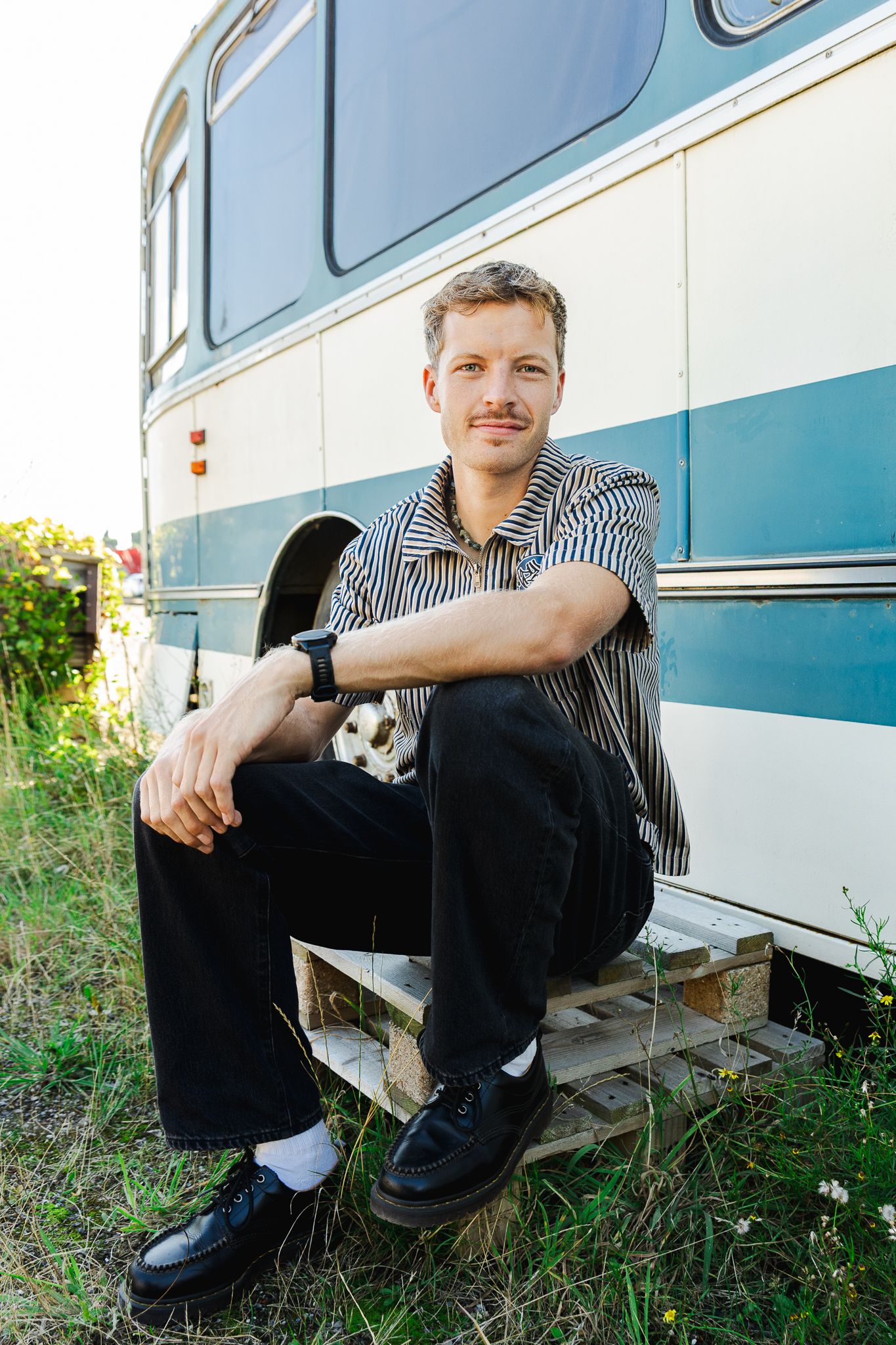 Man with mustache sitting on wooden pallet beside vintage blue and white bus outdoors on sunny day.