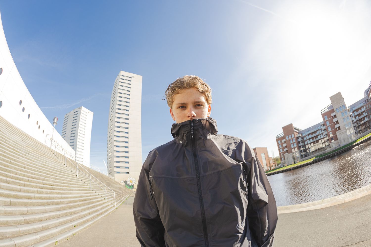 Young man in dark jacket standing outdoors near modern buildings and water on a sunny day.