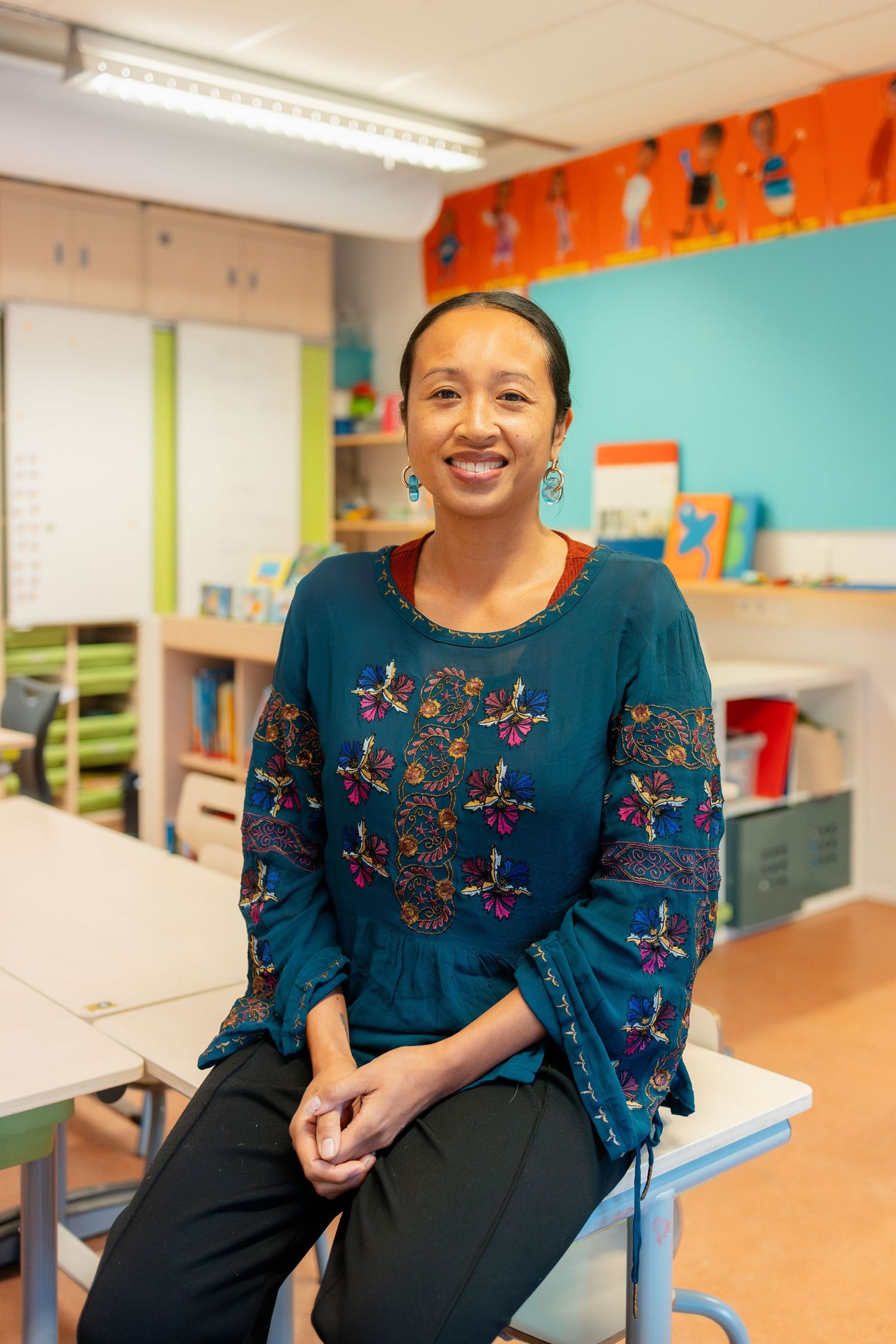A person wearing a navy blue dress with floral embroidery sits in a colorful classroom setting.