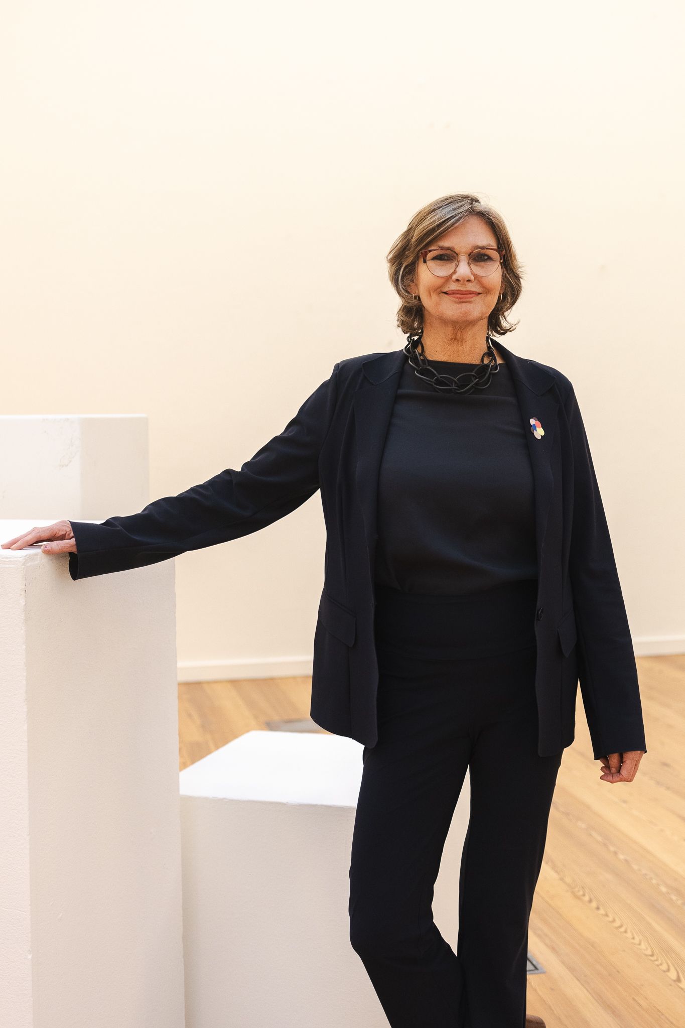A person wearing all black business attire stands next to a white pedestal in a gallery-like space with hardwood floors.