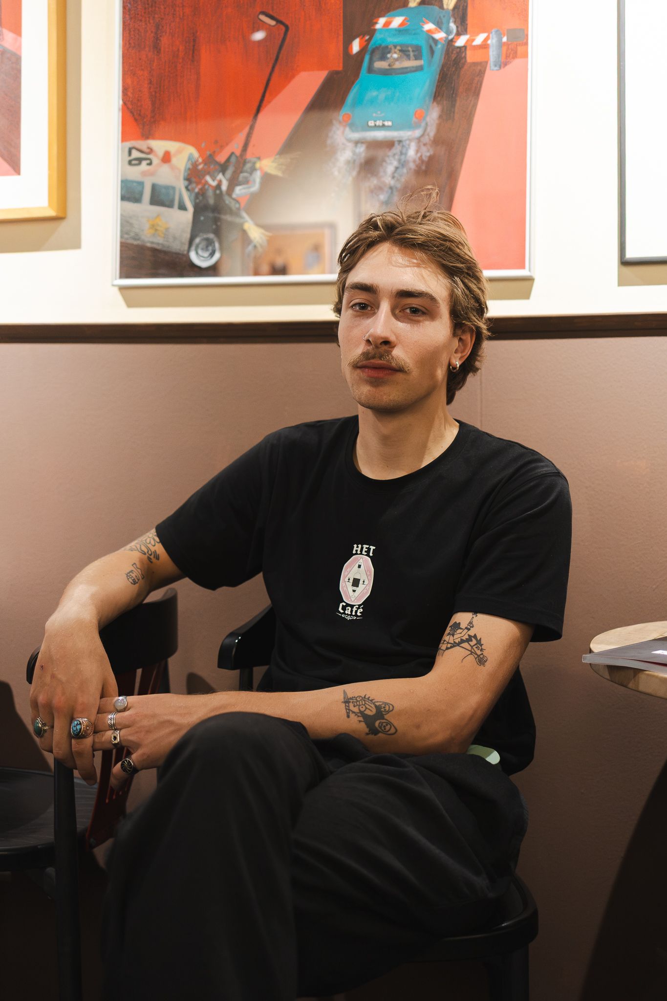 Young man with tattoos sitting in a chair wearing a black graphic t-shirt, with colorful artwork hanging on the wall behind him.