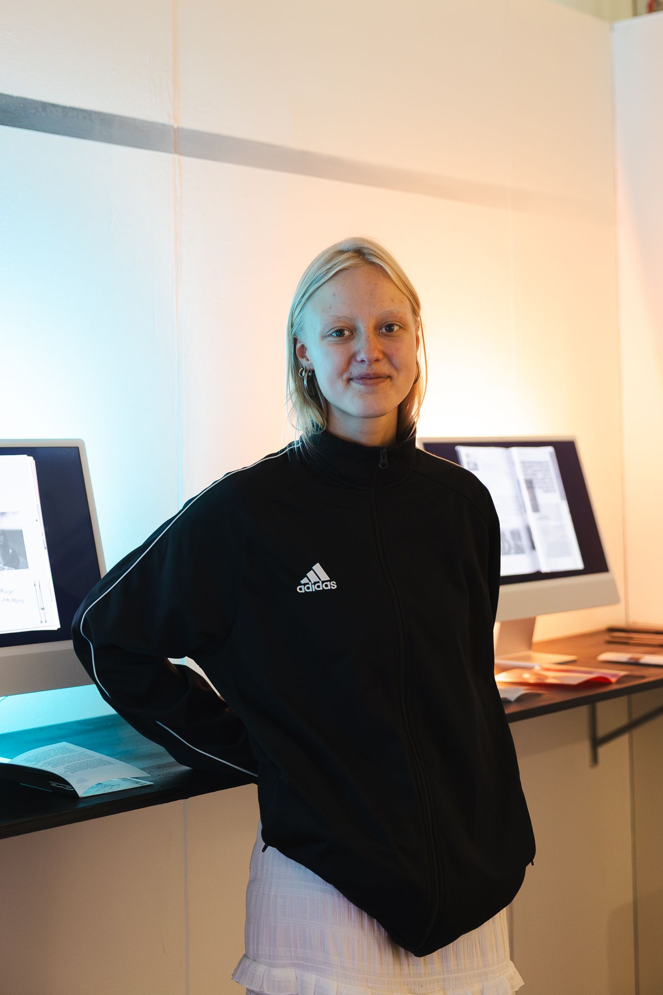 Young woman in black Adidas sweatshirt smiling indoors near display screens.