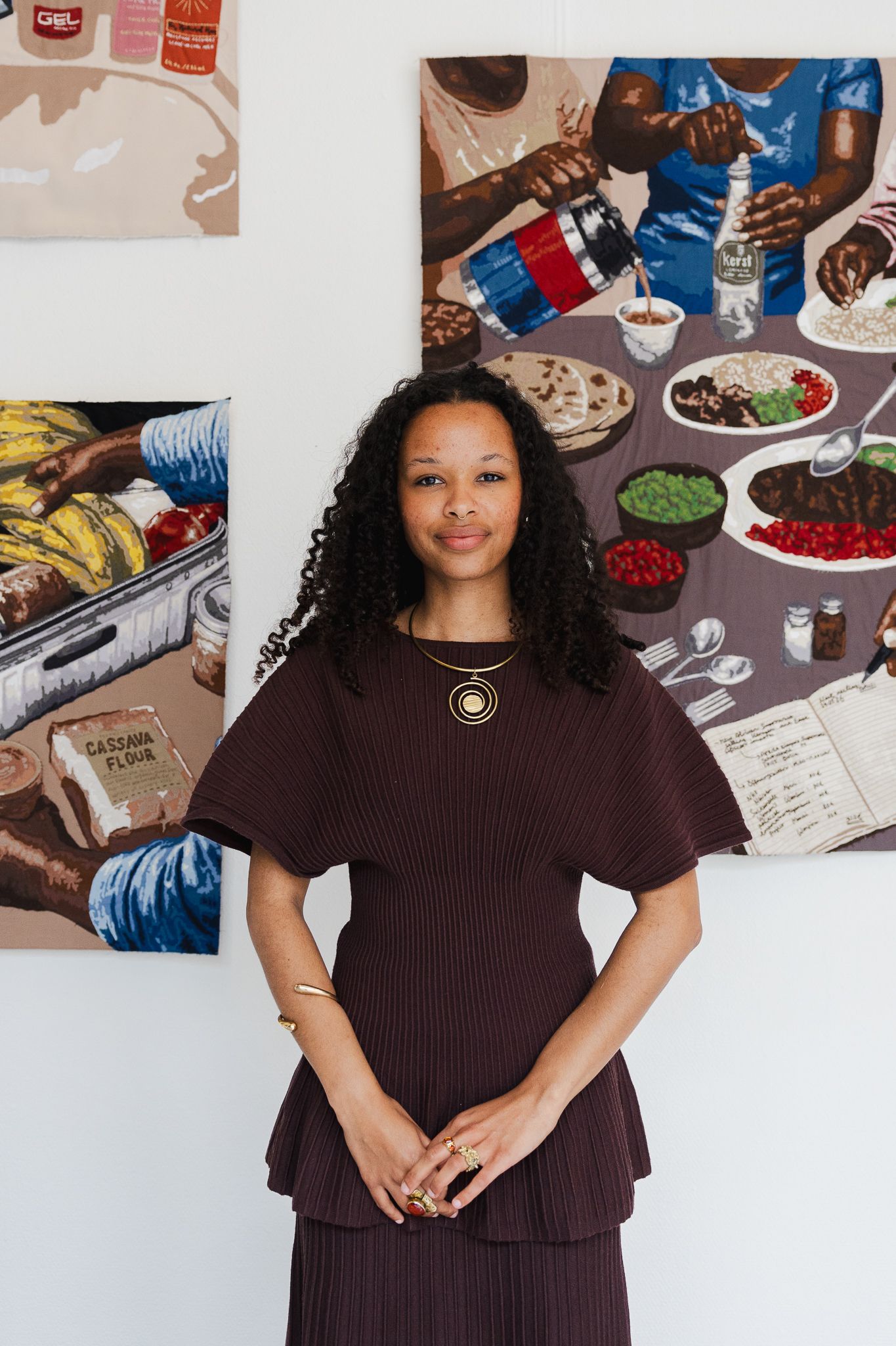 A woman in a dark burgundy ruffled top stands confidently in front of colorful food-themed paintings in an art gallery.