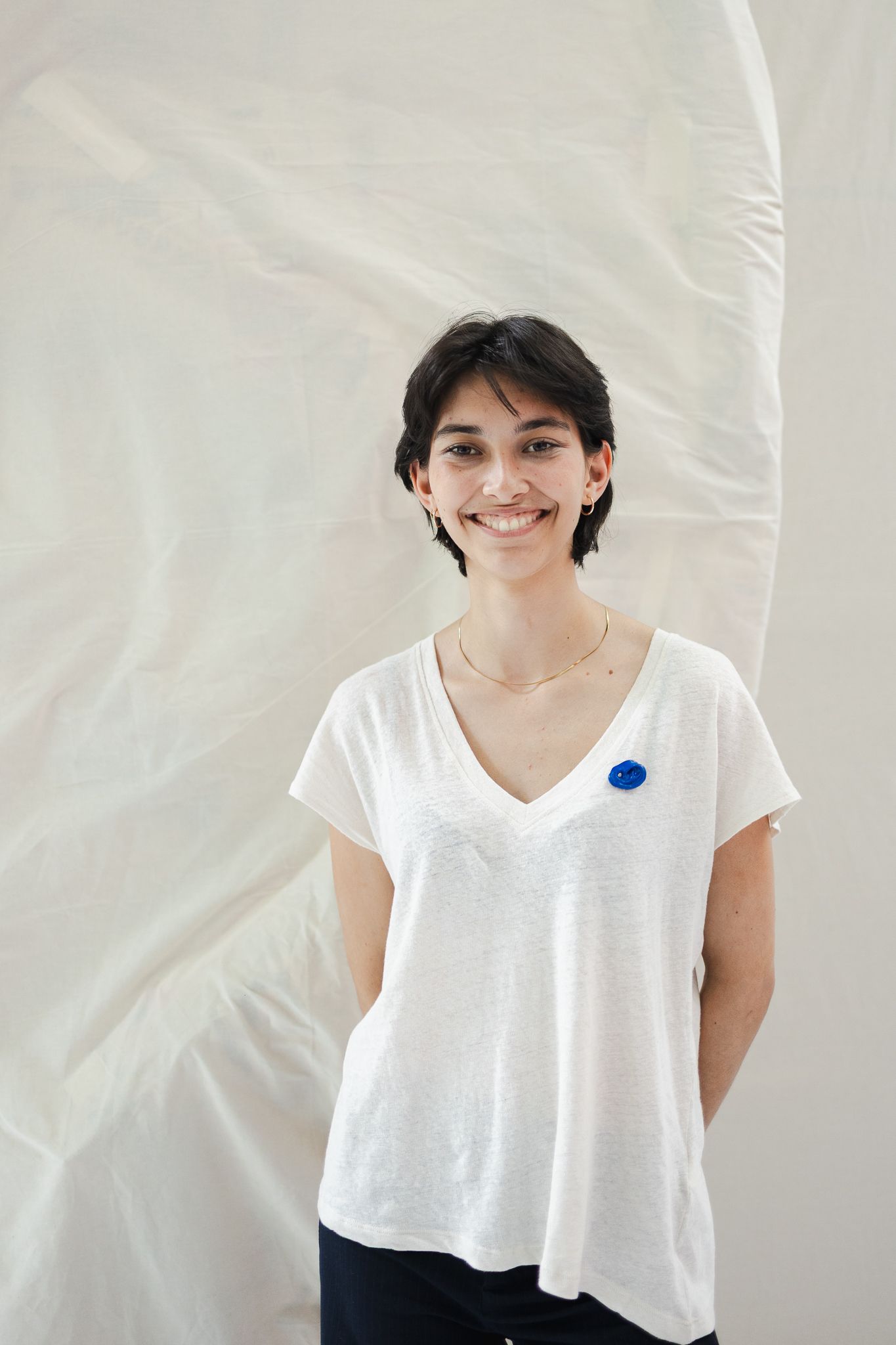 Smiling young woman with short dark hair wearing a white v-neck tee with a blue brooch, posing against a flowing white fabric backdrop.