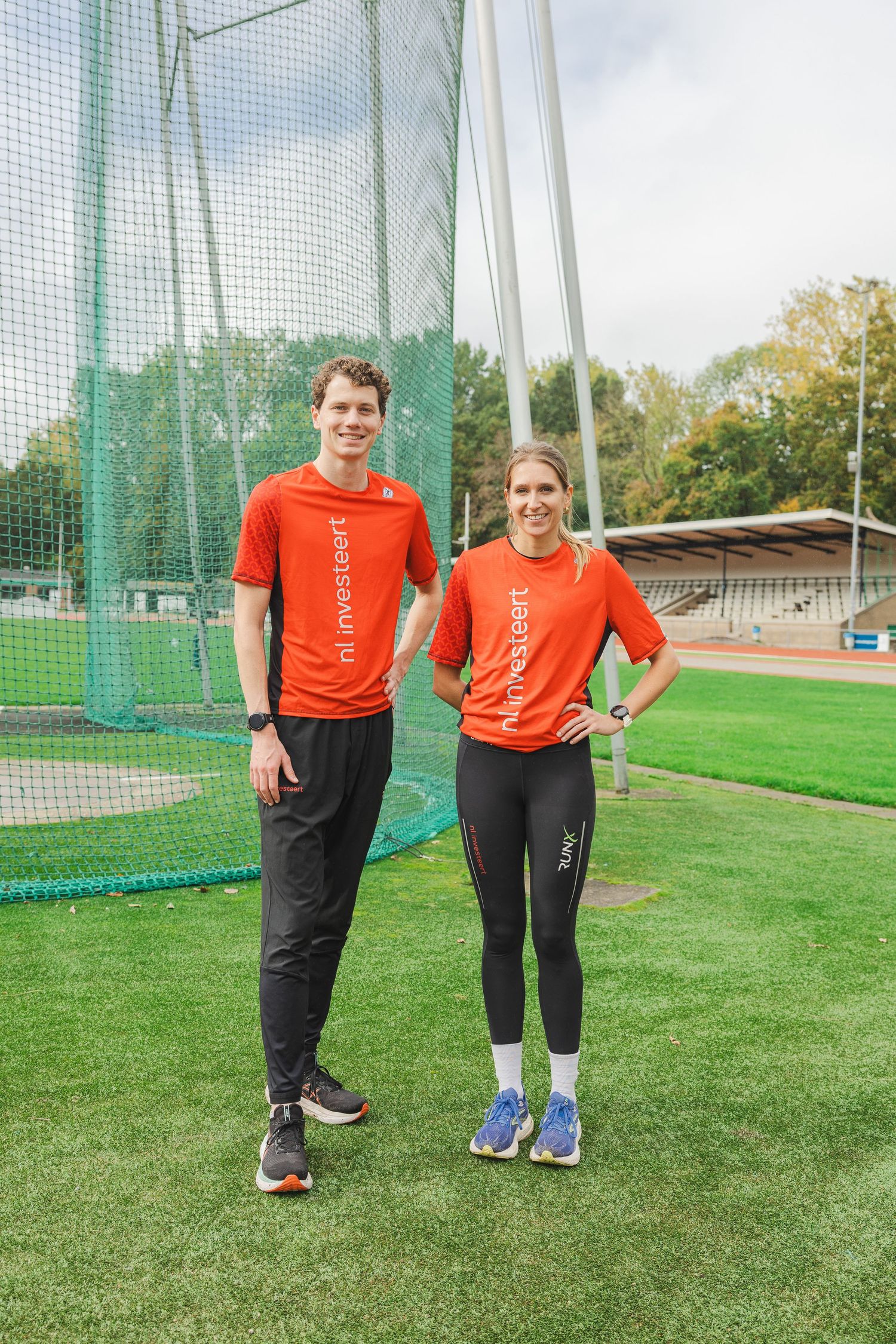 Two people wearing matching orange shirts stand together on a green athletic field with netting visible in the background.