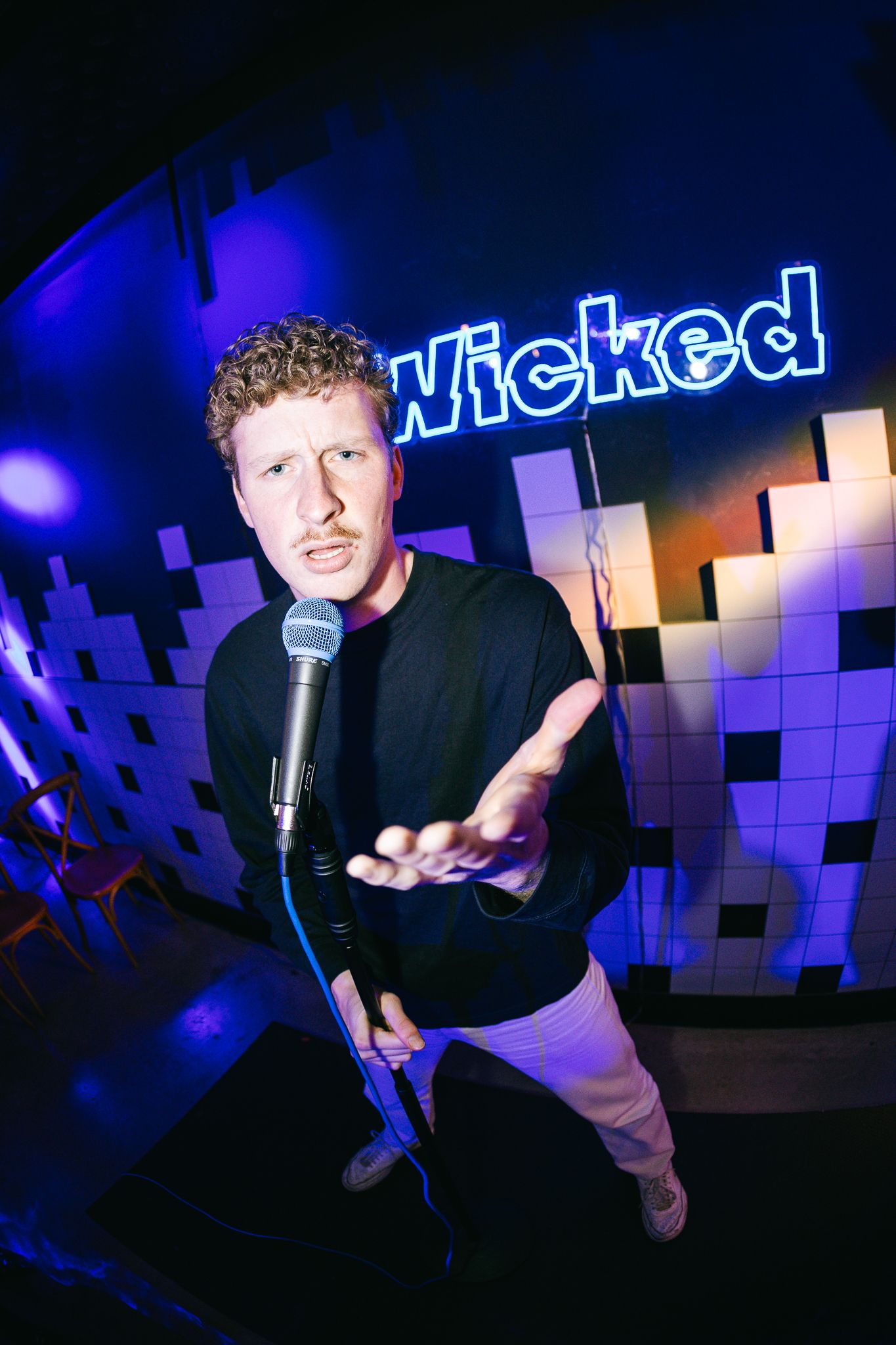 Man performing at mic stand under neon Wicked sign in blue-lit venue.