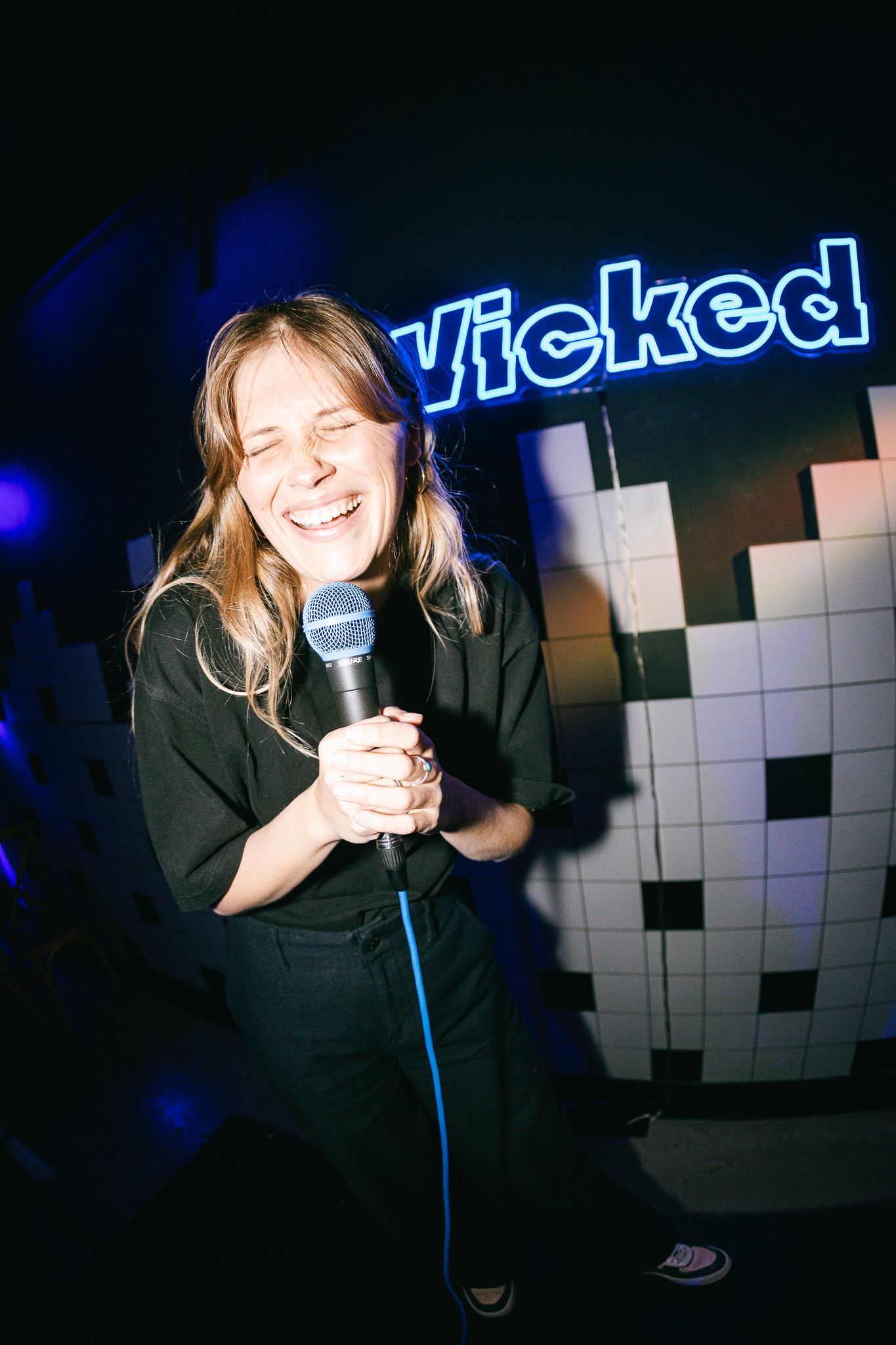 Woman laughing and singing into microphone at Wicked neon sign karaoke night event.