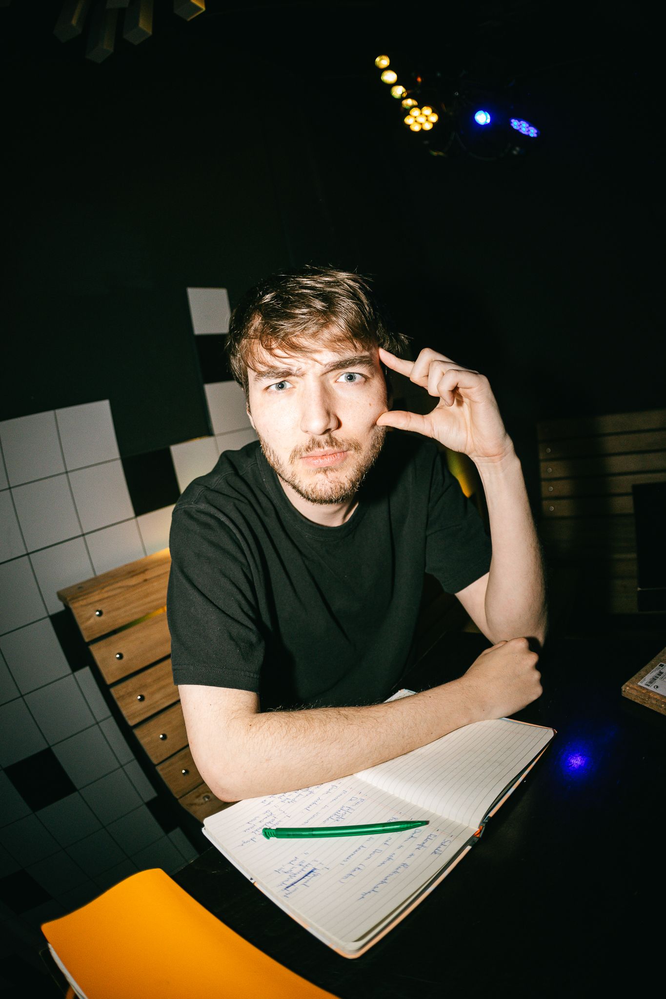 Young man in black t-shirt sits at desk with open notebook, hand raised to temple in thoughtful pose.