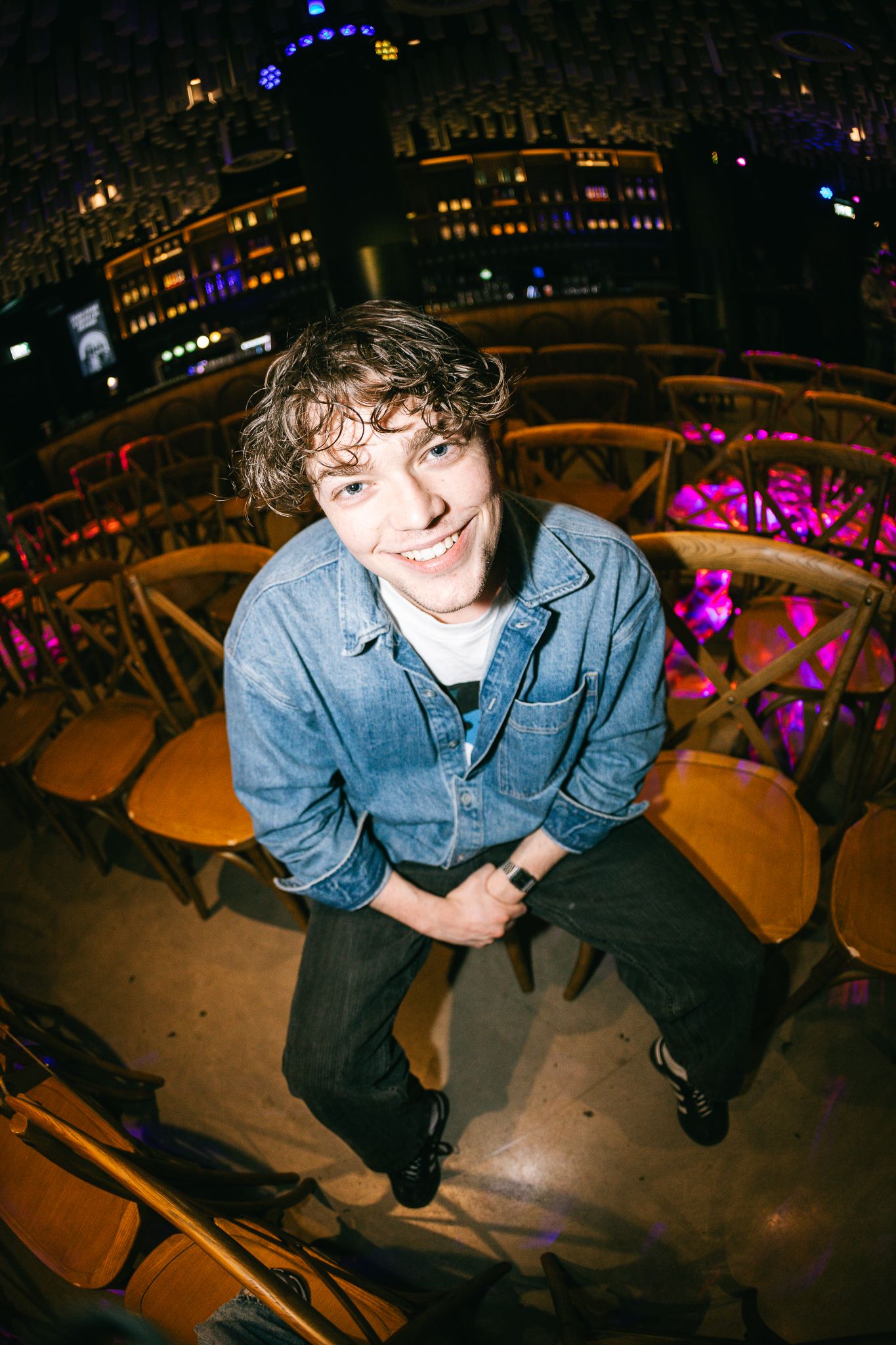 Young man with curly hair smiling in denim shirt, seated among wooden chairs in a colorful venue.