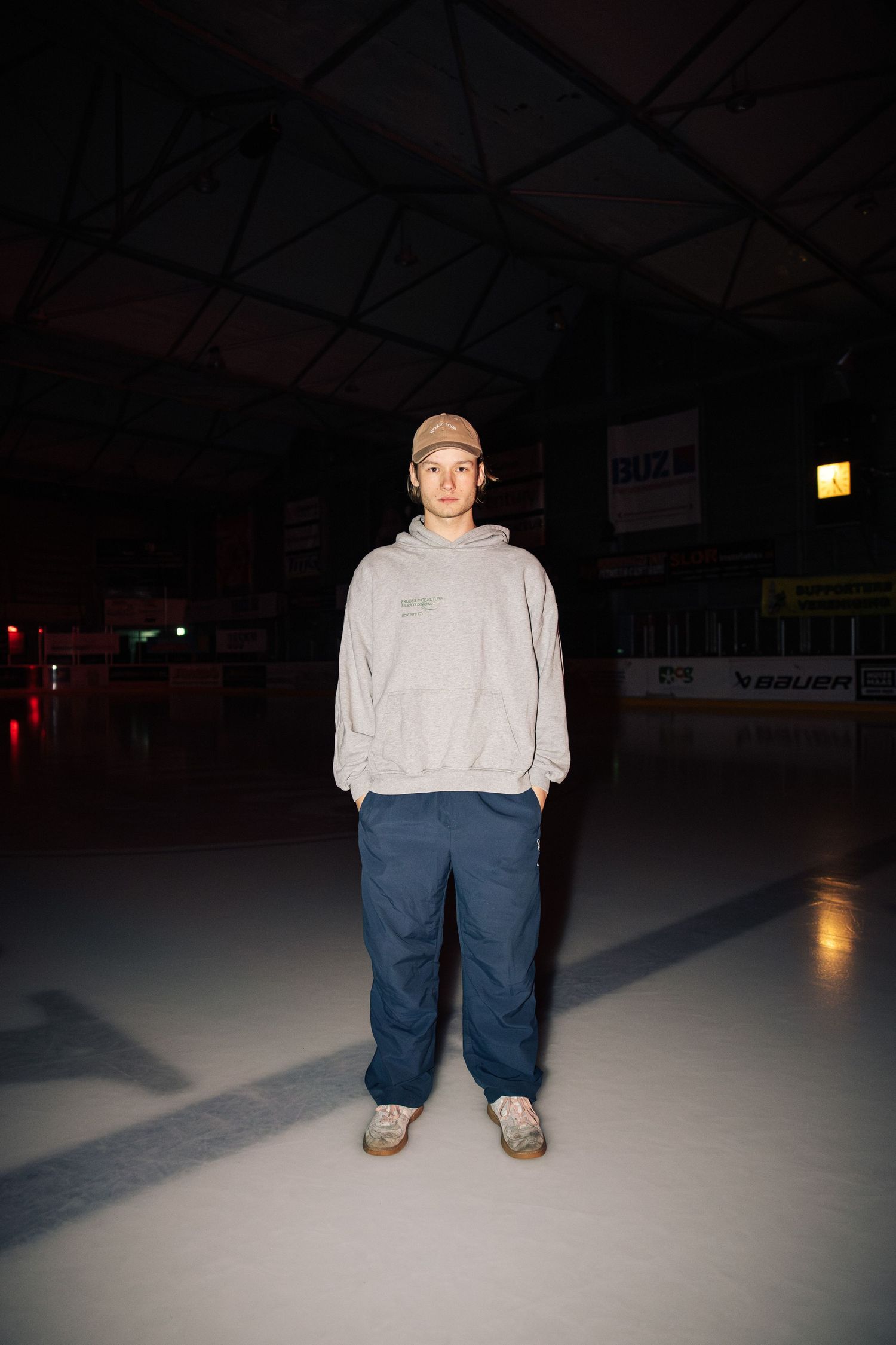Person standing alone on ice rink wearing grey hoodie and blue pants in dark indoor arena.