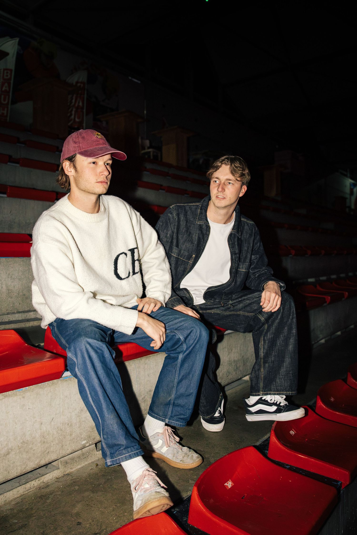 Two people sit on red bleachers wearing casual clothing including jeans and sweatshirts in a dimly lit setting.