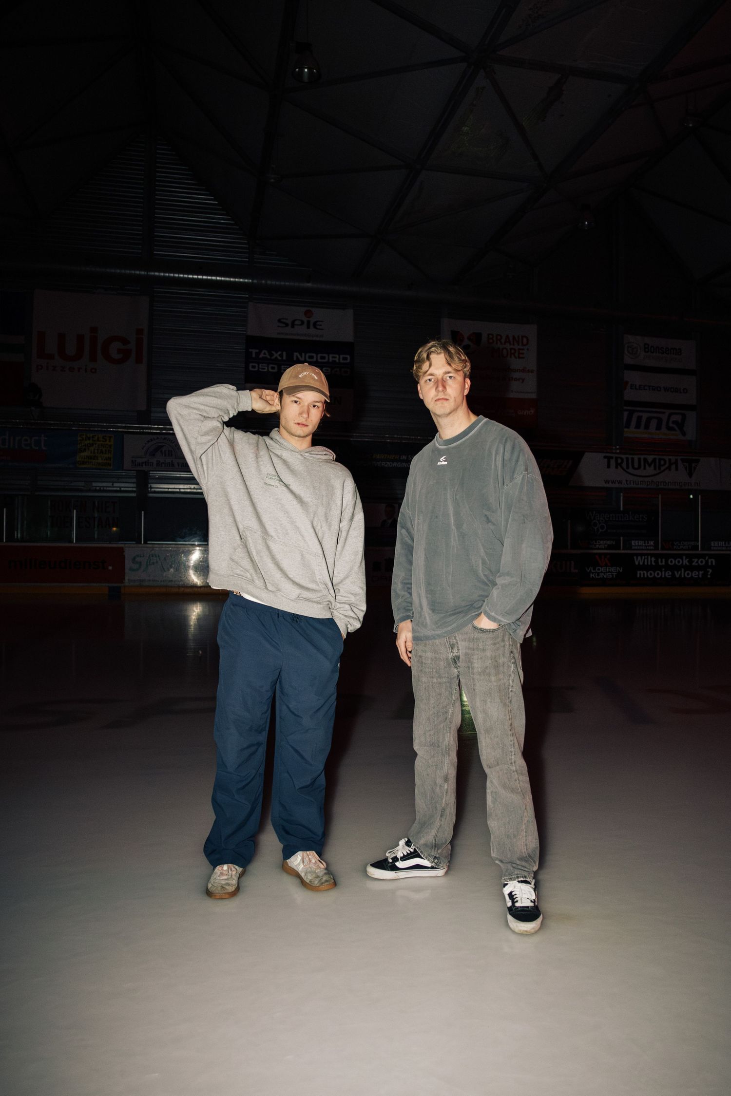 Two people sit together on the ground in a dimly lit indoor space wearing casual clothing.
