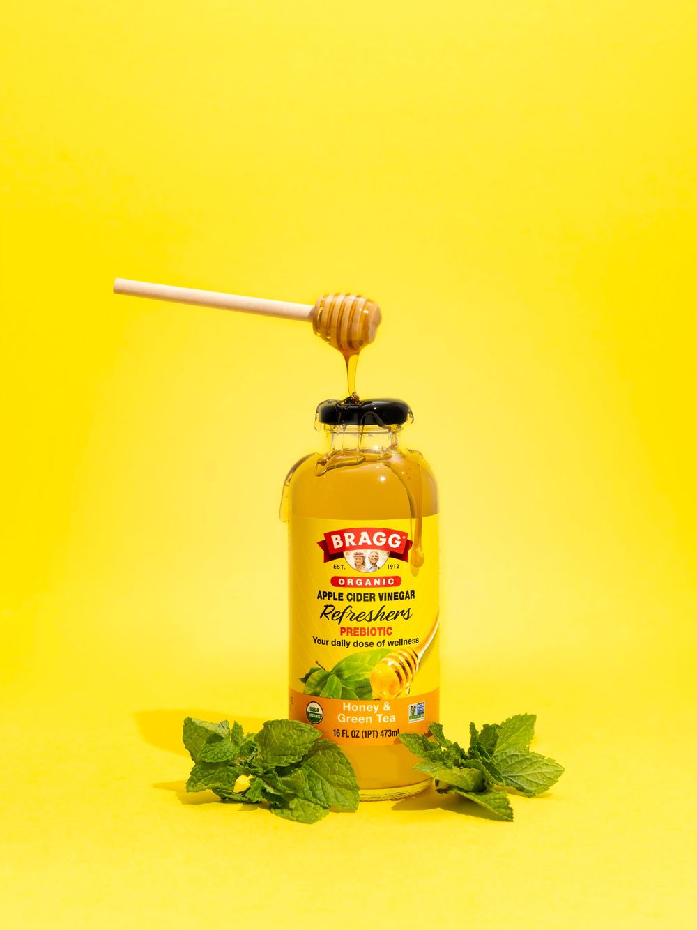 Honey dripping from wooden dipper onto bottle of Bragg apple cider vinegar with mint leaves on yellow background.