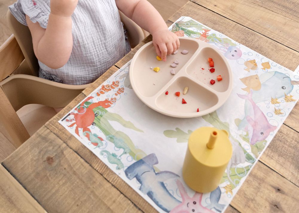 A toddler's dining setup with a sectioned pink plate and yellow cup on a colorful dinosaur placemat.