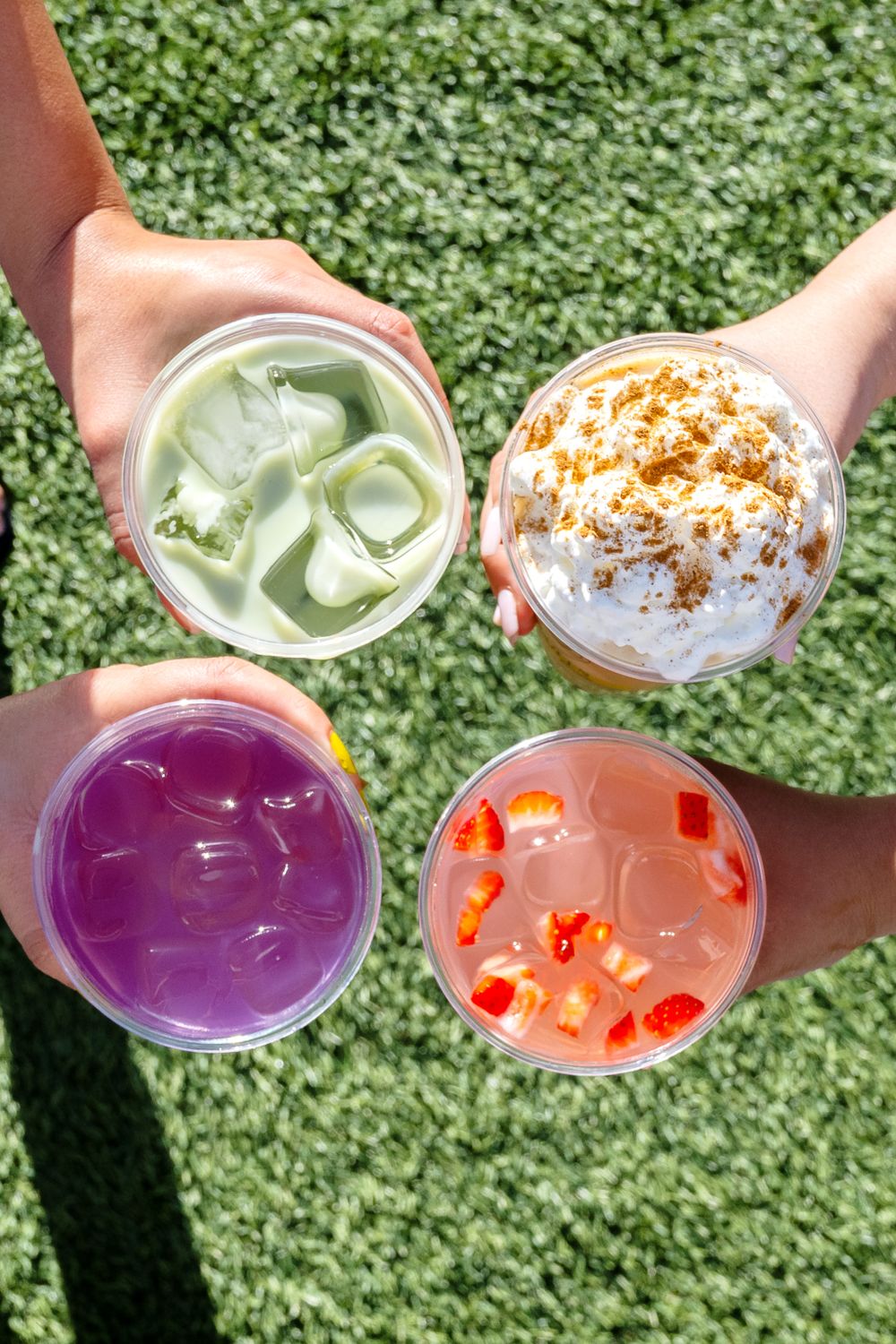 Overhead view of four colorful iced drinks on grass, including purple, green, and orange beverages.
