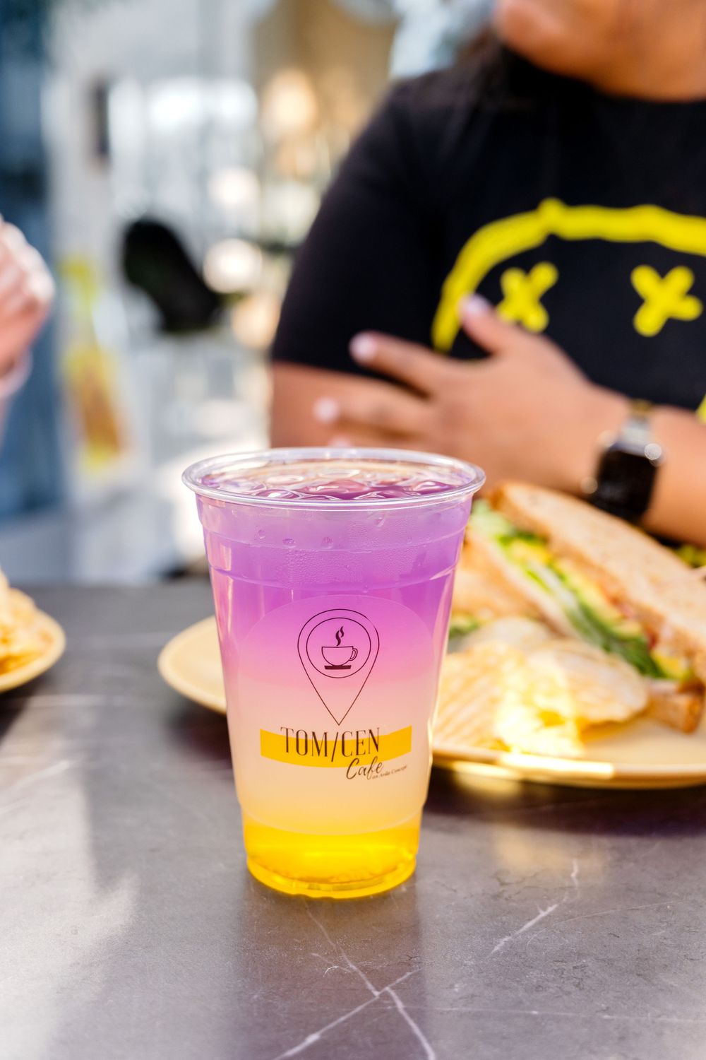 Gradient purple and yellow iced drink from Tom/Cen cafe with food in background.