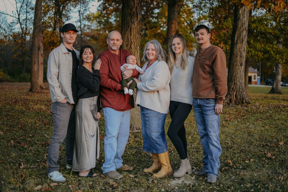 Multi-generational family of six poses outdoors among autumn trees, with grandparents holding a baby in the center.