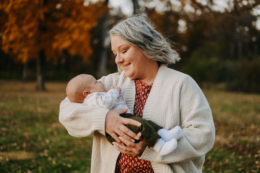 A smiling woman with gray hair holds a newborn baby outdoors in an autumn park with golden foliage.