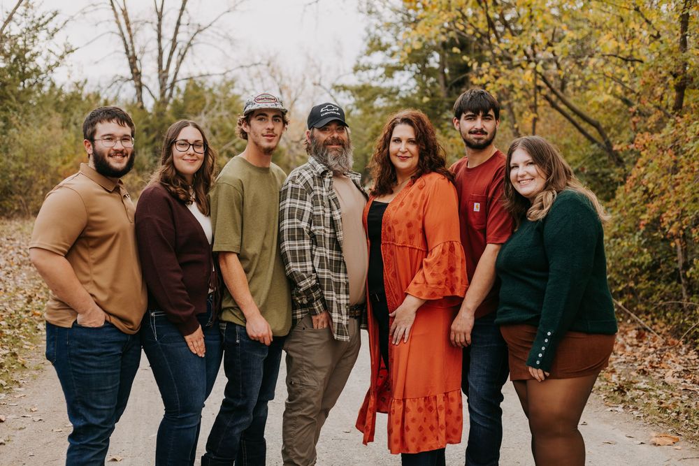 Family of seven smiling together outdoors among autumn foliage on a scenic wooded path.
