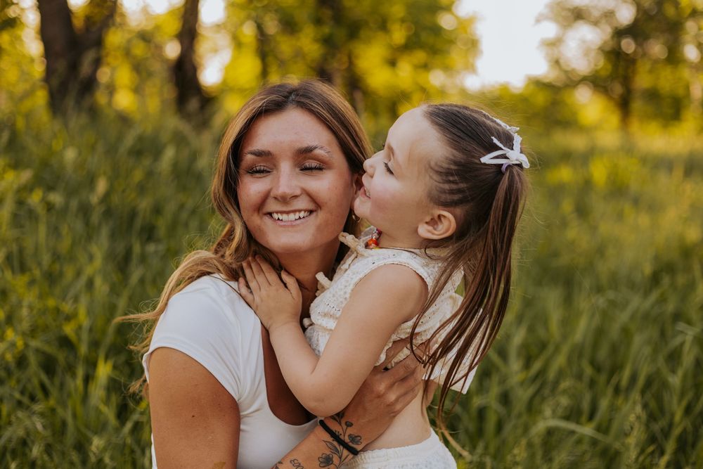 A young girl kisses her smiling mother on the cheek outdoors in a sunny green field.