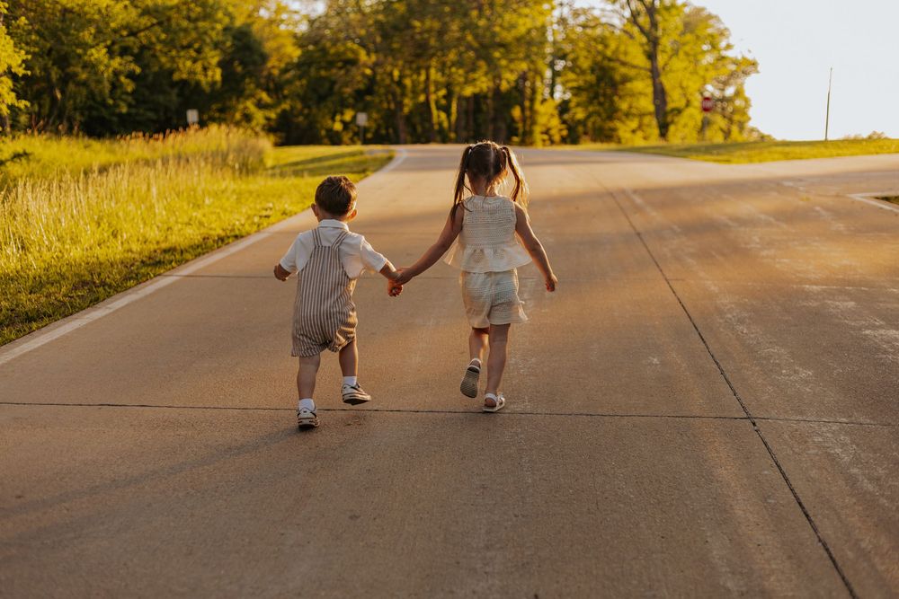Two young children holding hands while walking down a sunlit road surrounded by green trees at golden hour.