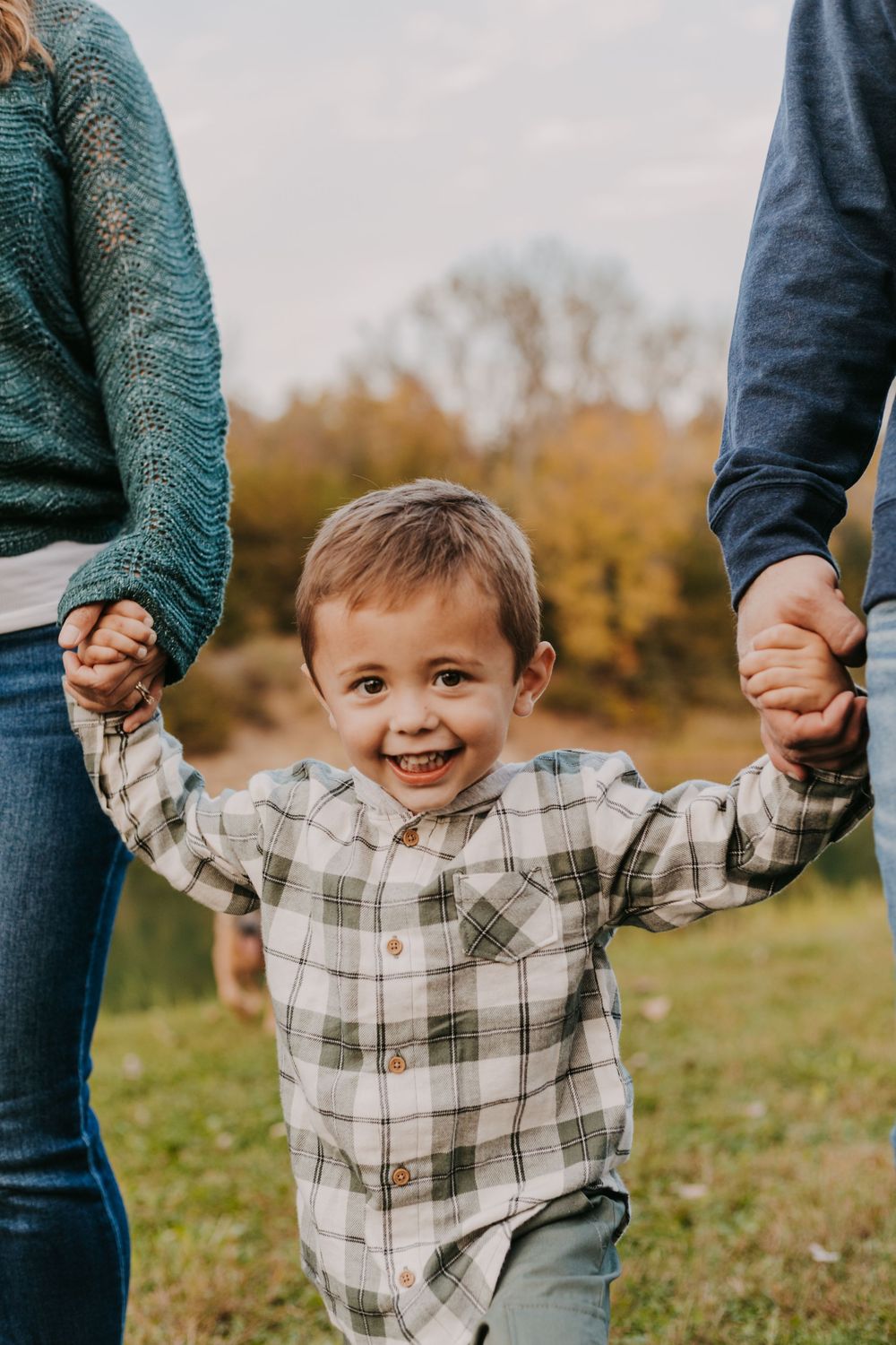 A happy toddler boy in a plaid shirt holds his parents hands outdoors during a fall family photo session.