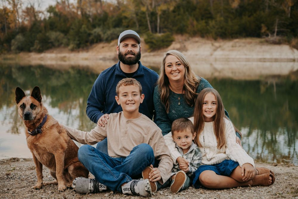 Family of five with a dog posing outdoors near a reflective lake surrounded by autumn trees.