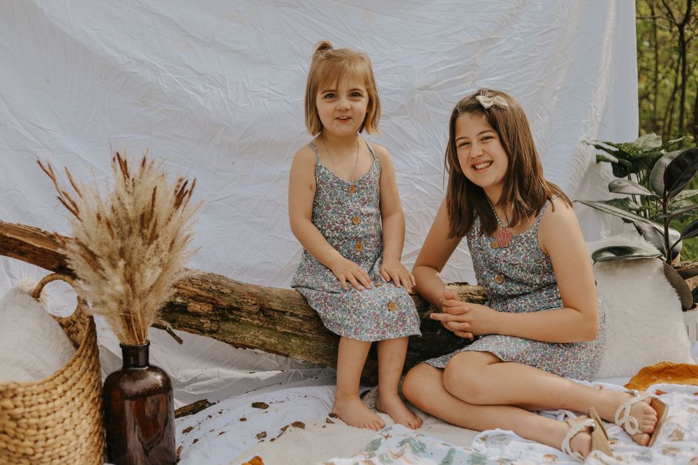 Two people in matching floral dresses sit together on a white blanket with boho decor.