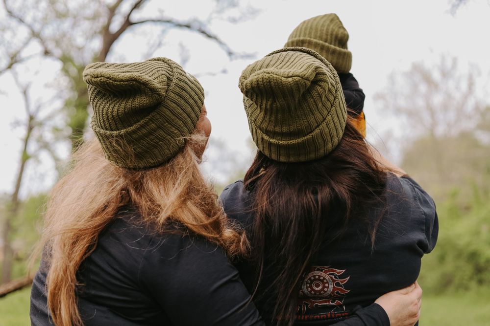 Two friends wearing matching olive green beanies sit together outdoors on a sunny day.