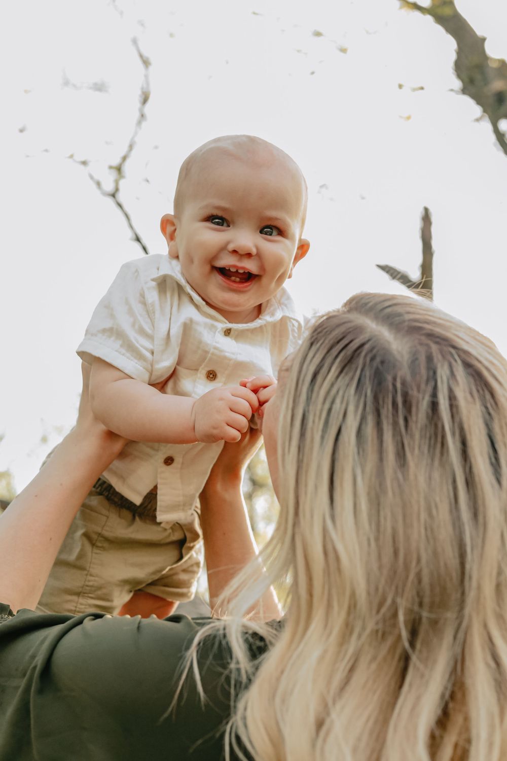 A baby in beige overalls smiles joyfully while being lifted in the air outdoors.