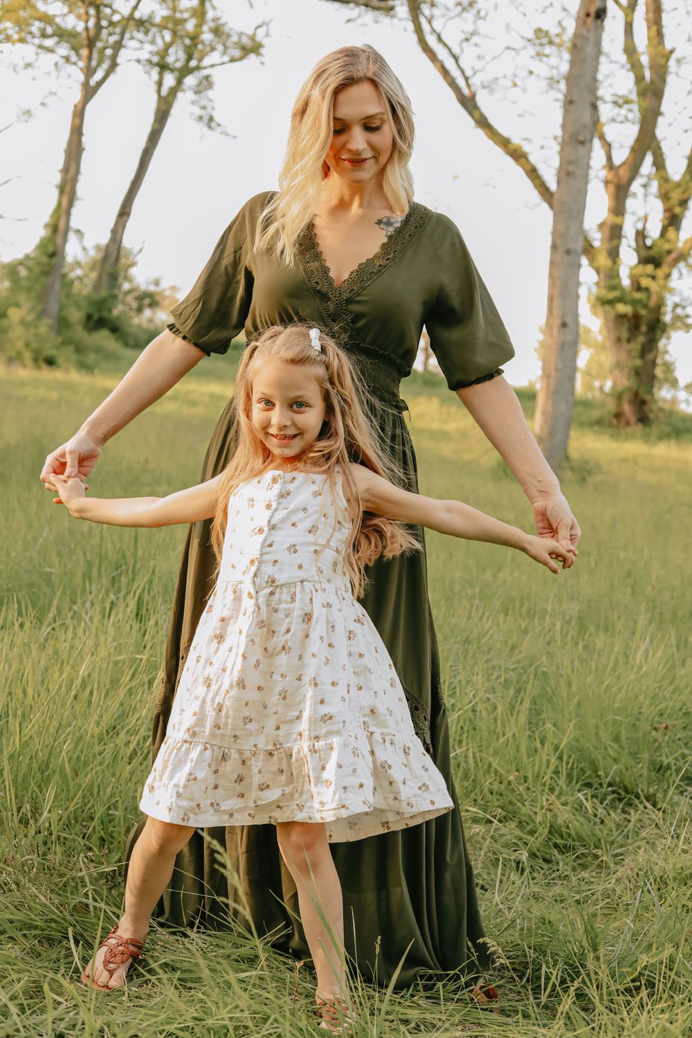 Woman in olive dress and child in white floral dress hold hands while standing in tall grass.