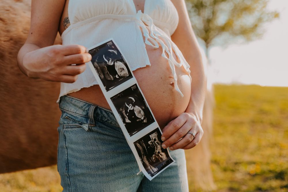 Pregnant woman holding ultrasound photos against her bare belly outdoors in golden hour light, wearing white top and jeans.