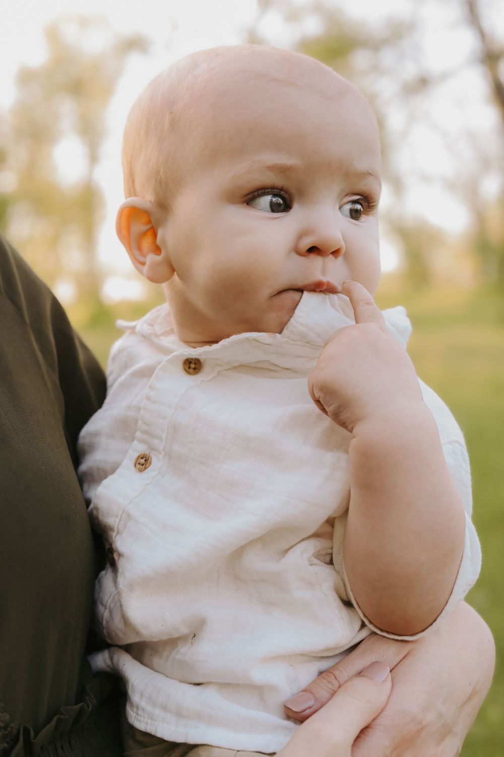 Baby in white button-up shirt with fingers in mouth held against dark clothing in outdoor setting.