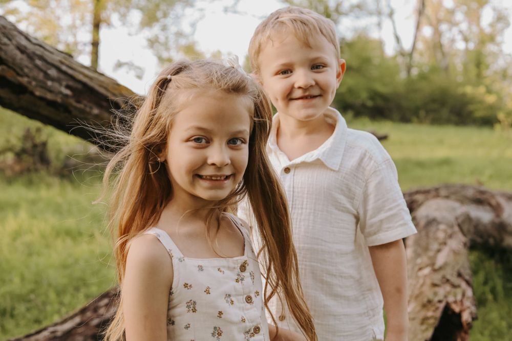 Two young children smiling while posing near a fallen log in a natural outdoor setting.
