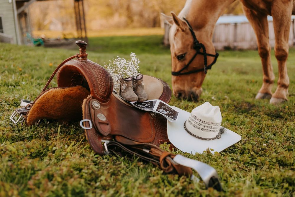 Western saddle and cowboy hats on grass with a grazing palomino horse in the background.