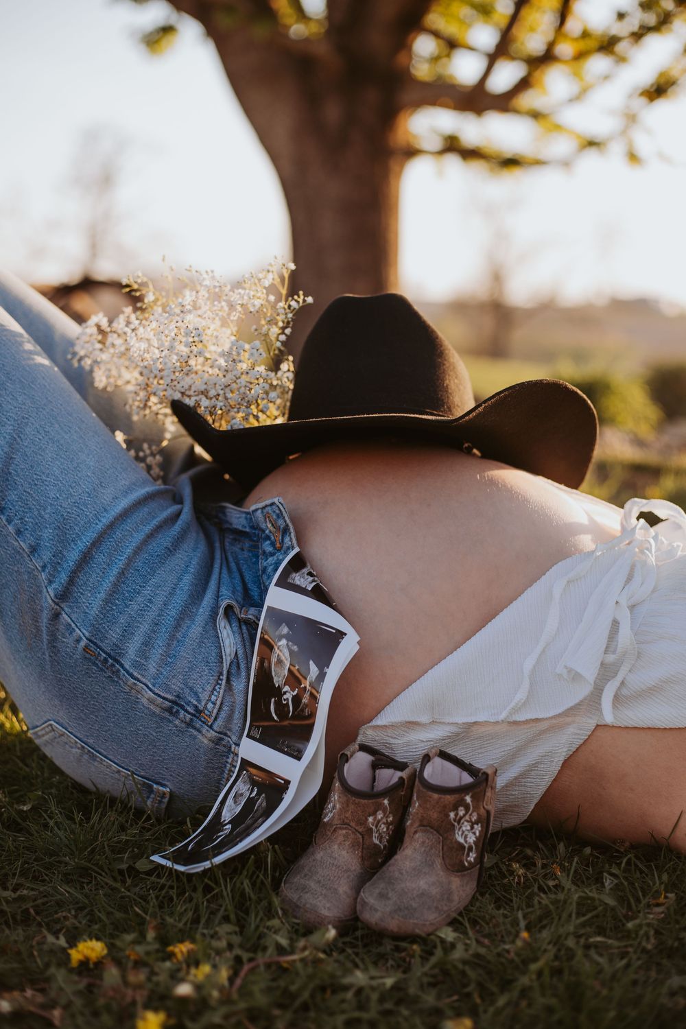 Pregnant woman lying on grass with cowboy hat on belly, baby boots, ultrasound photos, and wildflowers in golden sunlight.