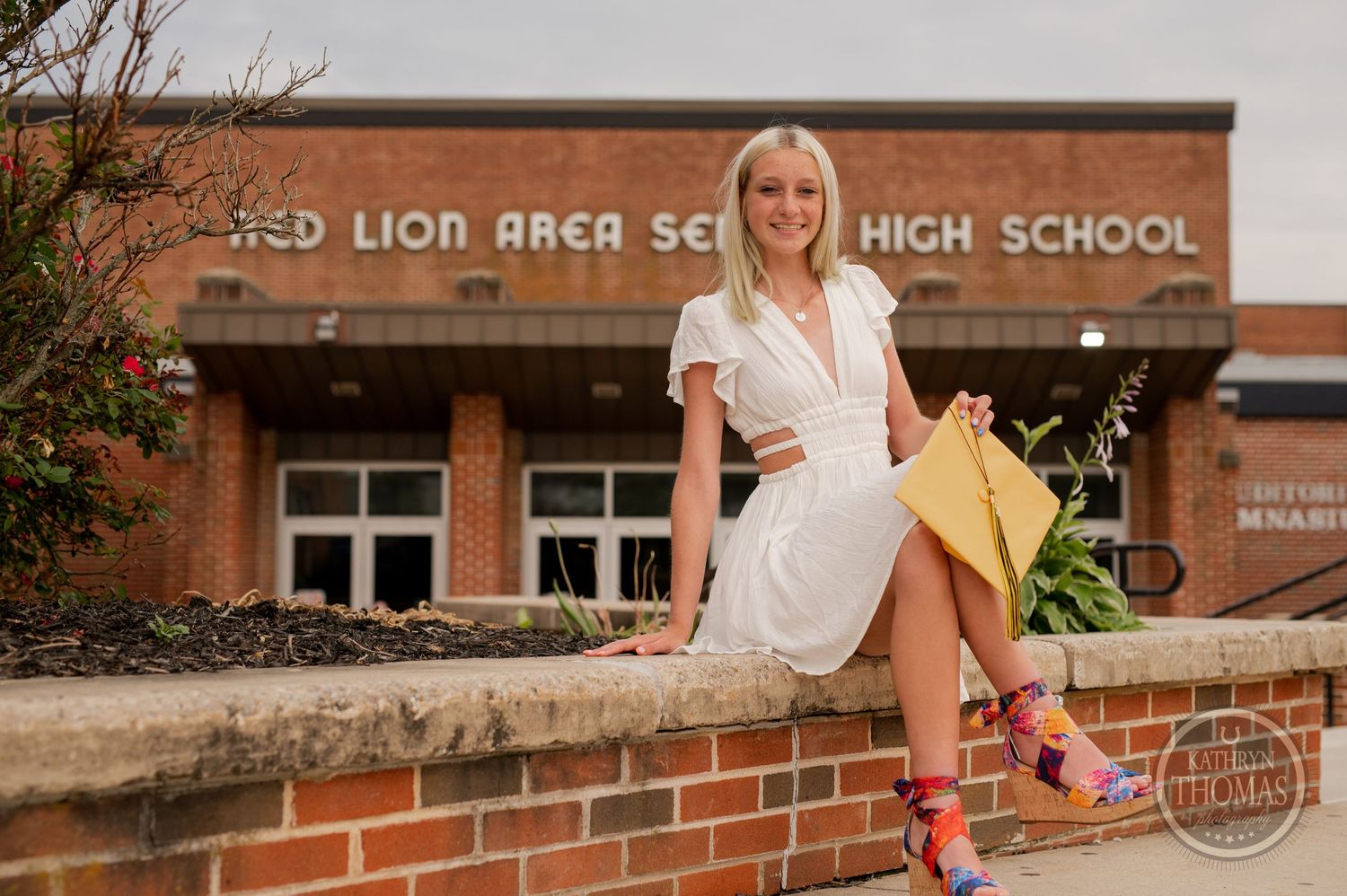 Graduate in white dress sits casually on brick wall outside Red Lion Area Senior High.