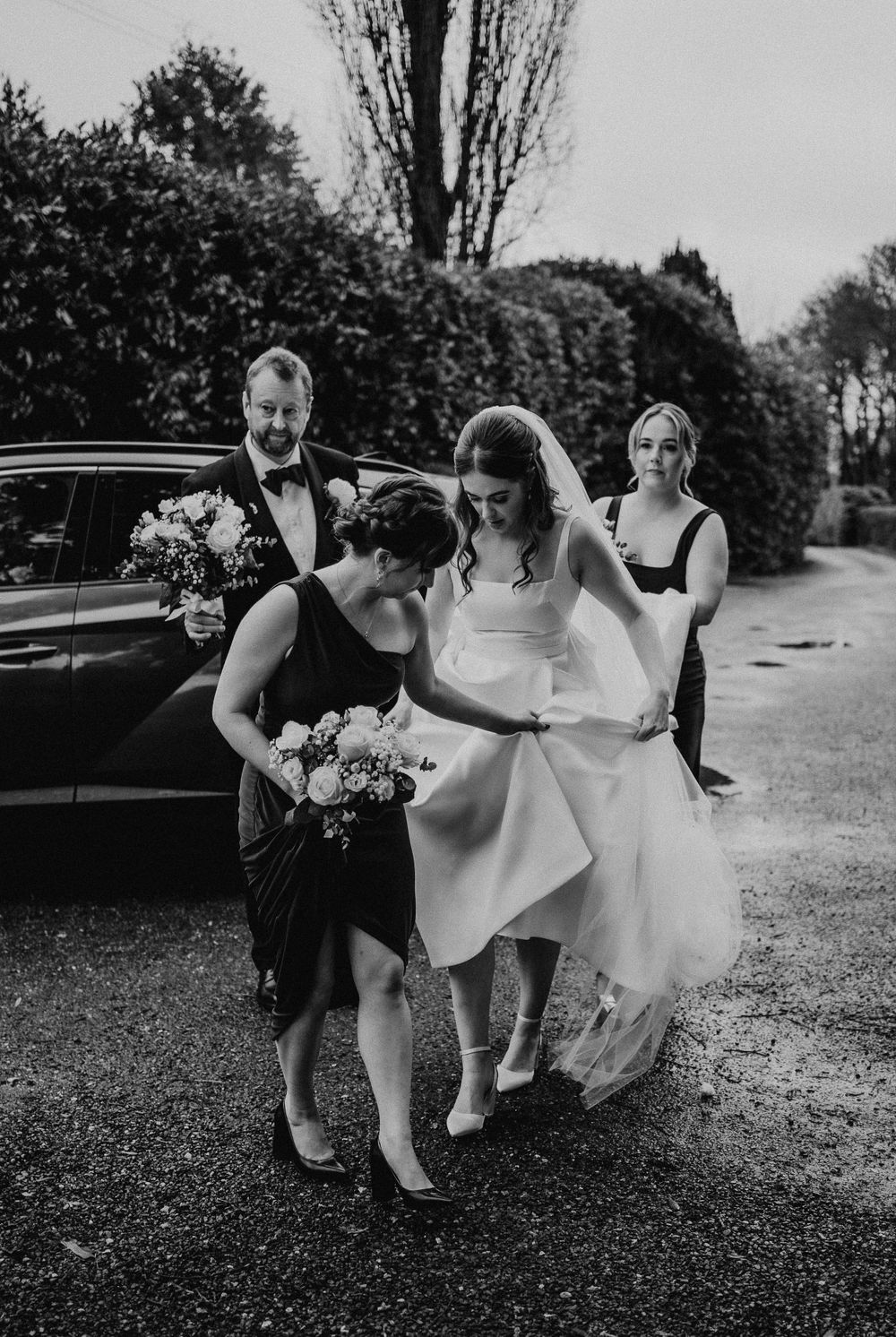 Wedding party in black dresses helps adjust bride's white gown outdoors in black and white photo.