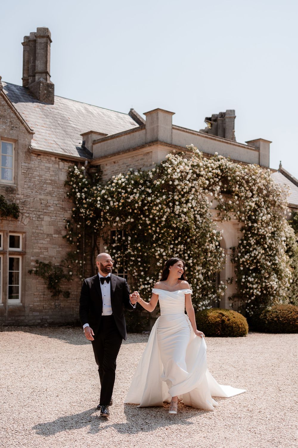 A couple in formal wedding attire walks along a gravel path beside a stone building.